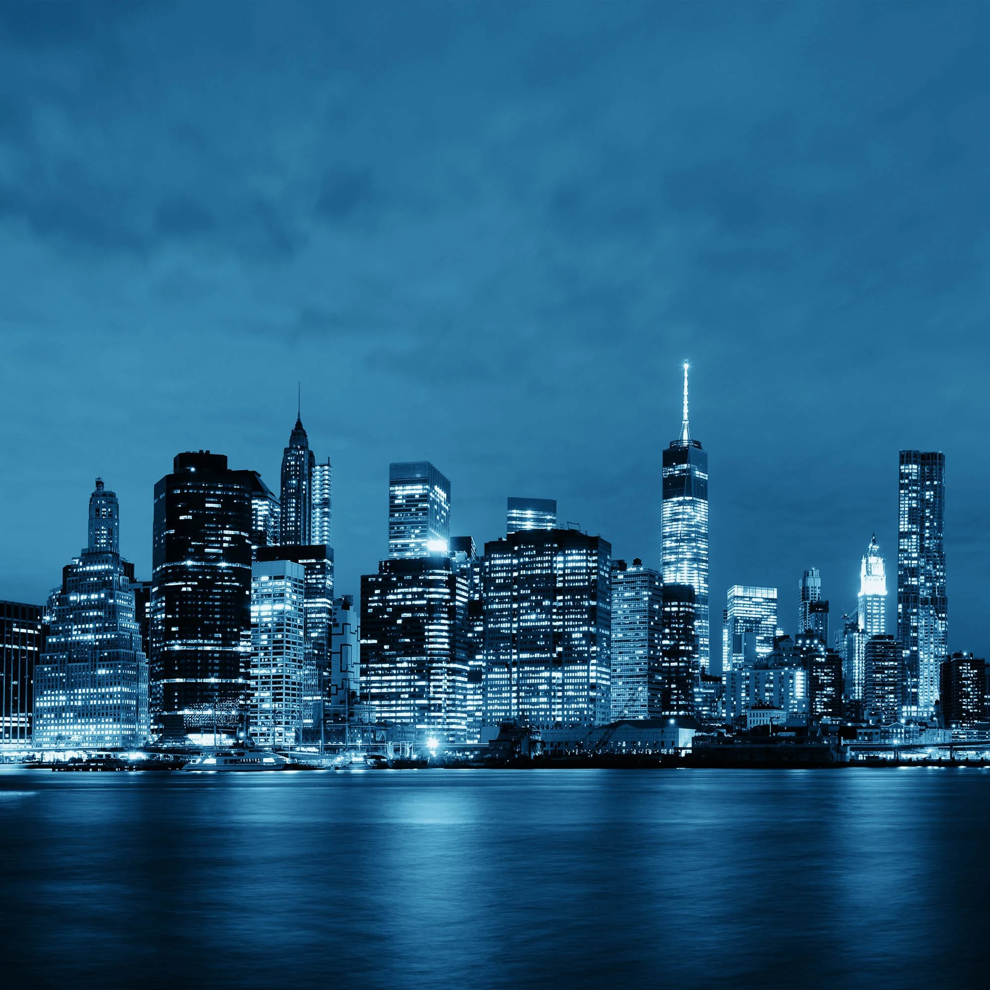 Nighttime view of New York City skyline with tall illuminated skyscrapers, including the Empire State Building and One World Trade Center, reflecting on the river in the foreground.