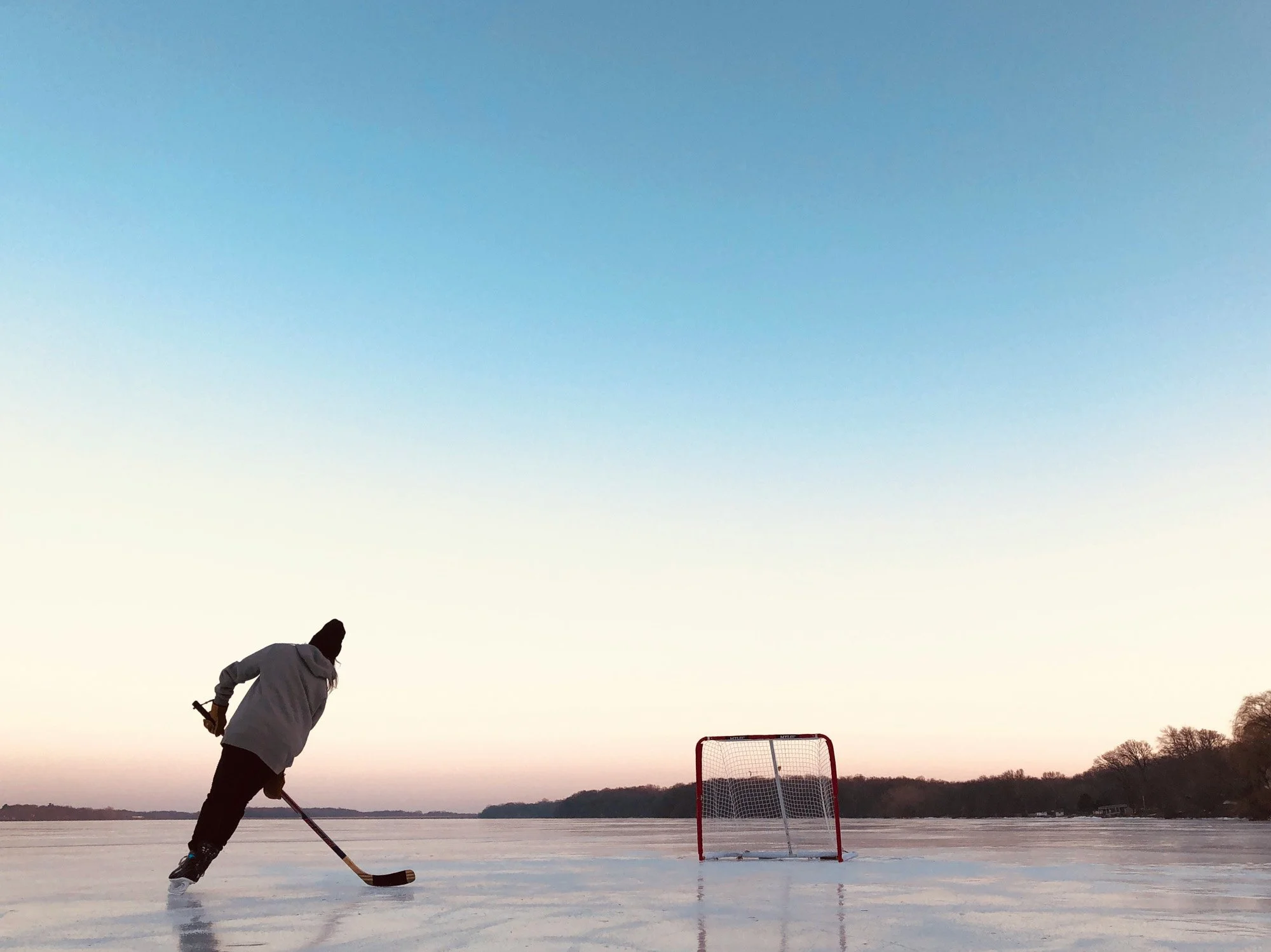Person playing hockey on outdoor ice rink near a hockey goal at sunset or sunrise.