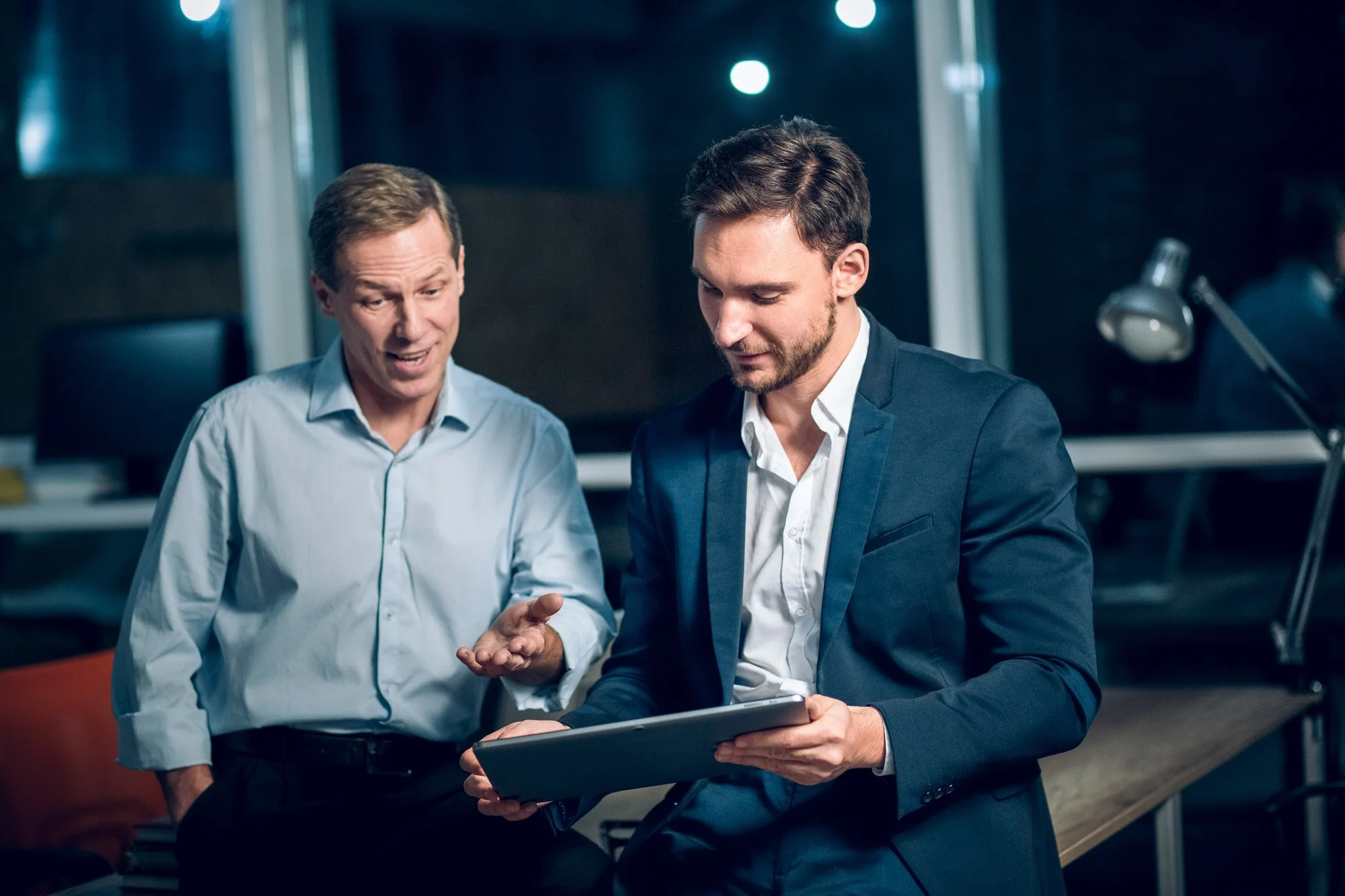 Two men in business attire looking at a tablet in an office setting.
