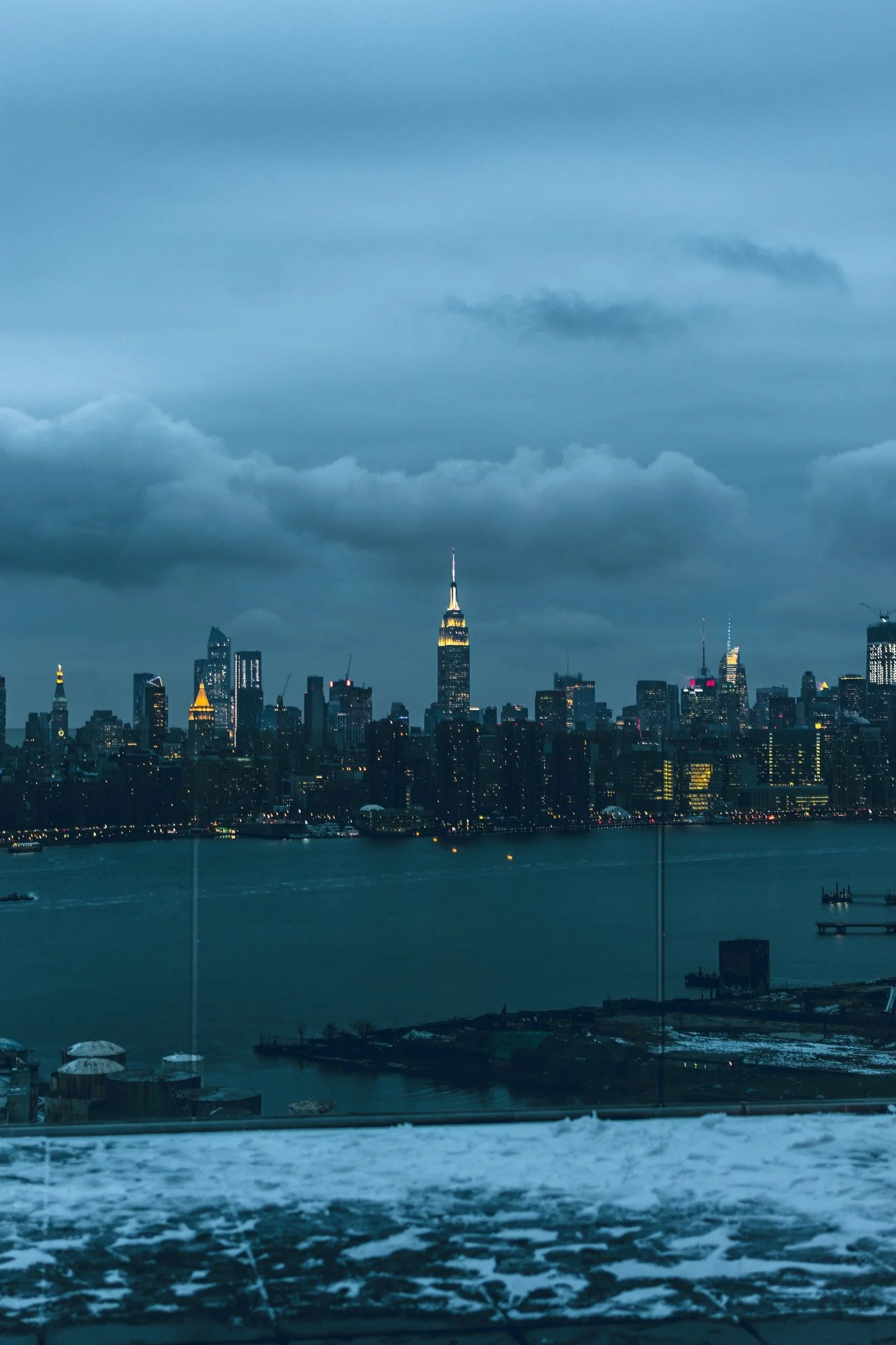 Nighttime view of the New York City skyline with the Empire State Building illuminated, across a river, under a cloudy sky.
