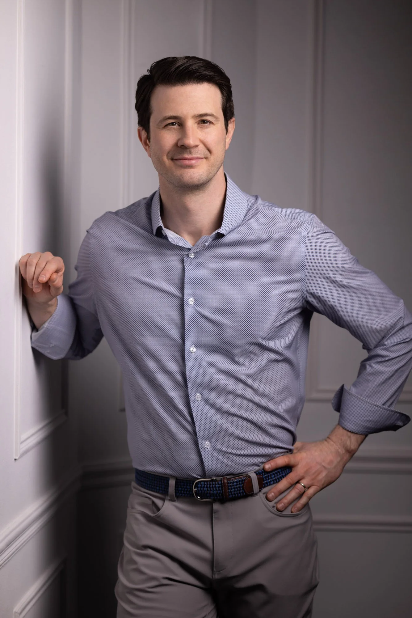 A man in a light blue patterned shirt and beige pants leans against a wall, smiling at the camera.