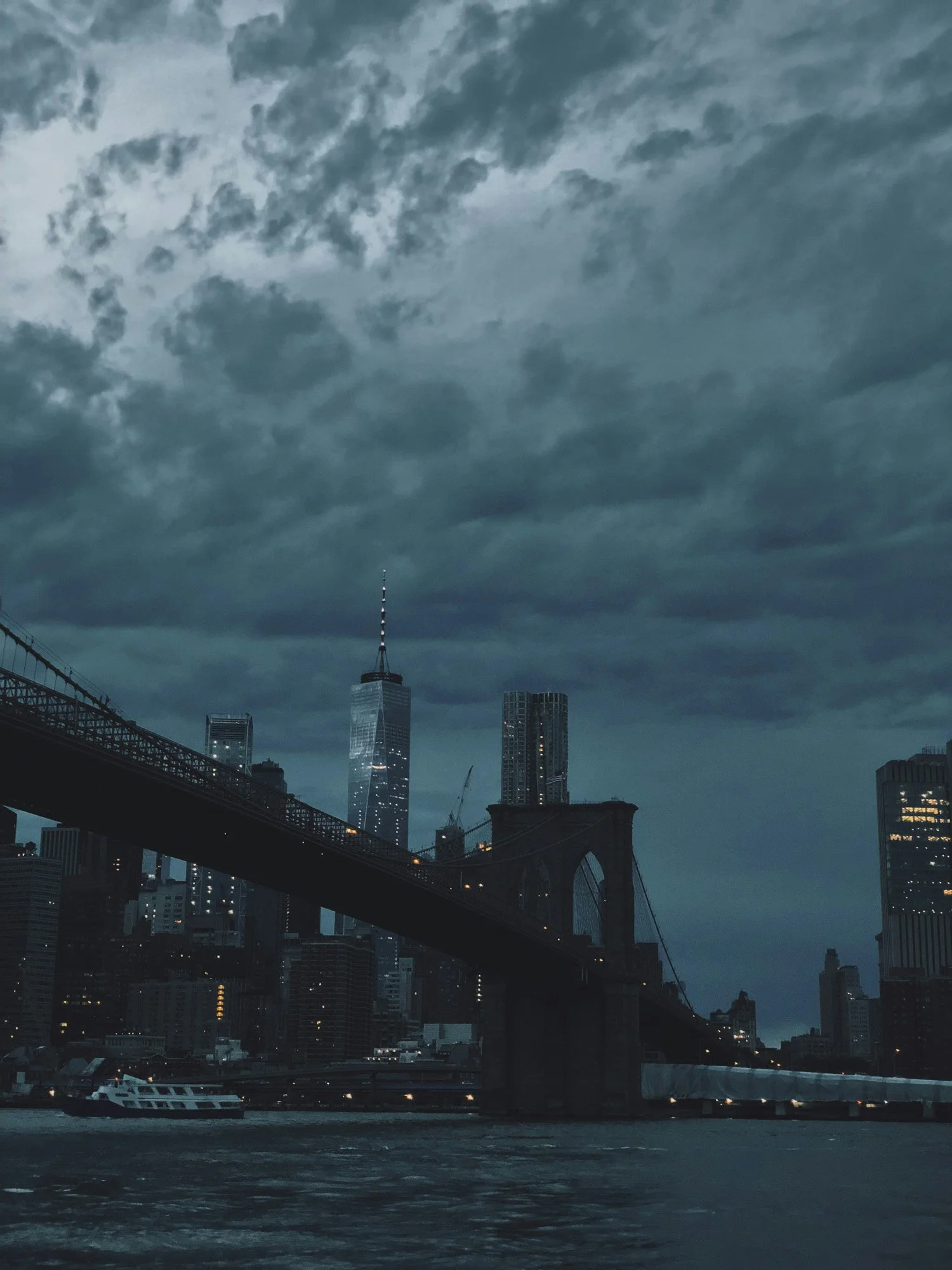 Nighttime view of the New York City skyline with the Brooklyn Bridge in the foreground and One World Trade Center under cloudy skies.