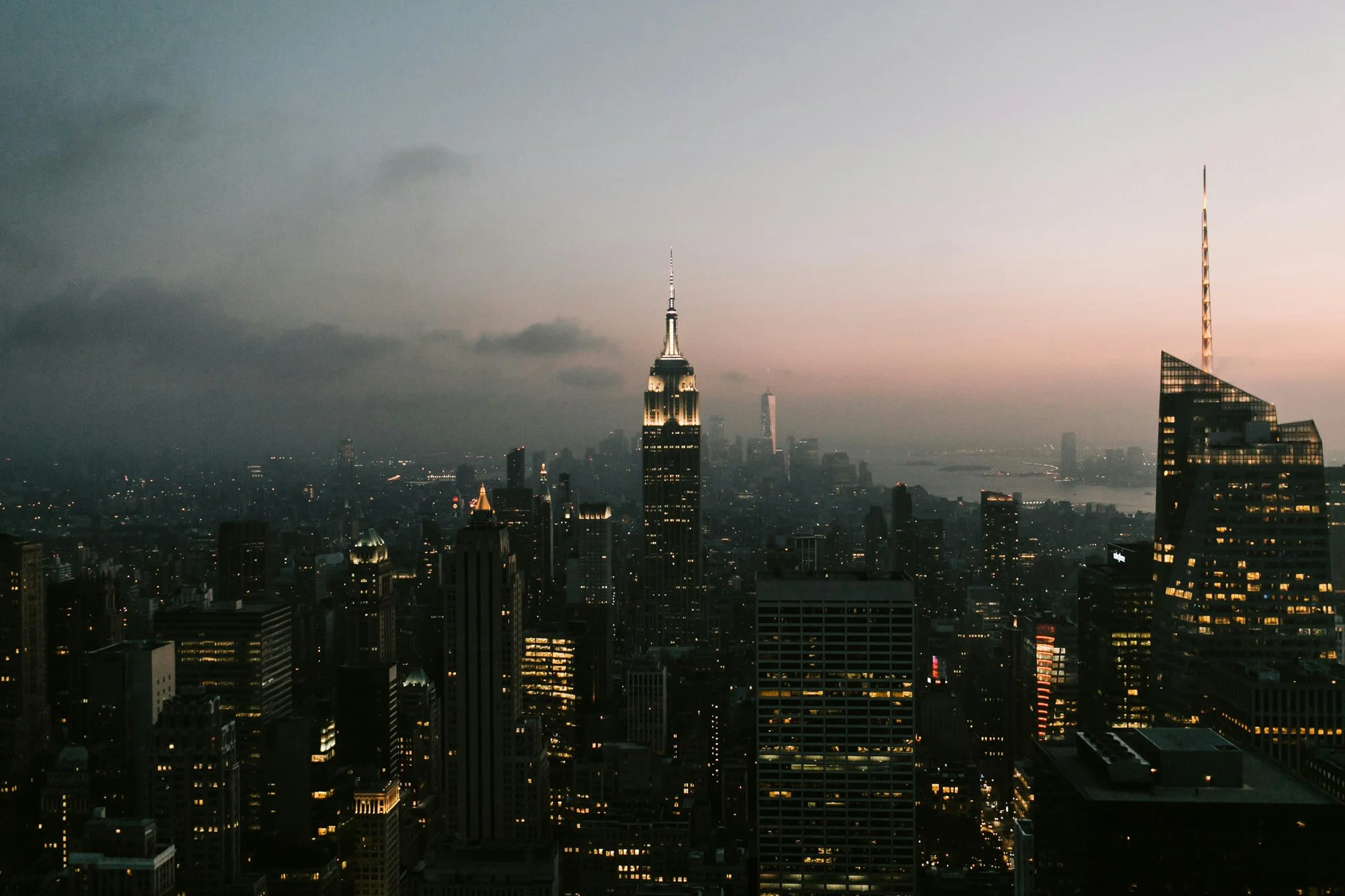 Dark cityscape of New York City skyline at dusk with illuminated skyscrapers, including the Empire State Building and One World Trade Center, with a cloudy sky and faint pink sunset glow.