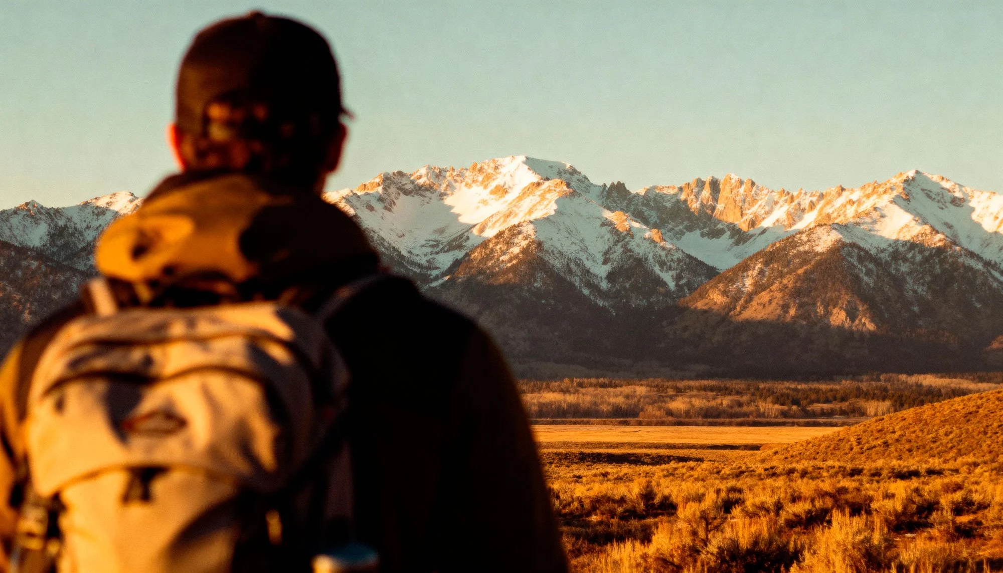 A person wearing hiking gear and a hat, facing a mountain range with snow-capped peaks, in a landscape at sunset or sunrise.