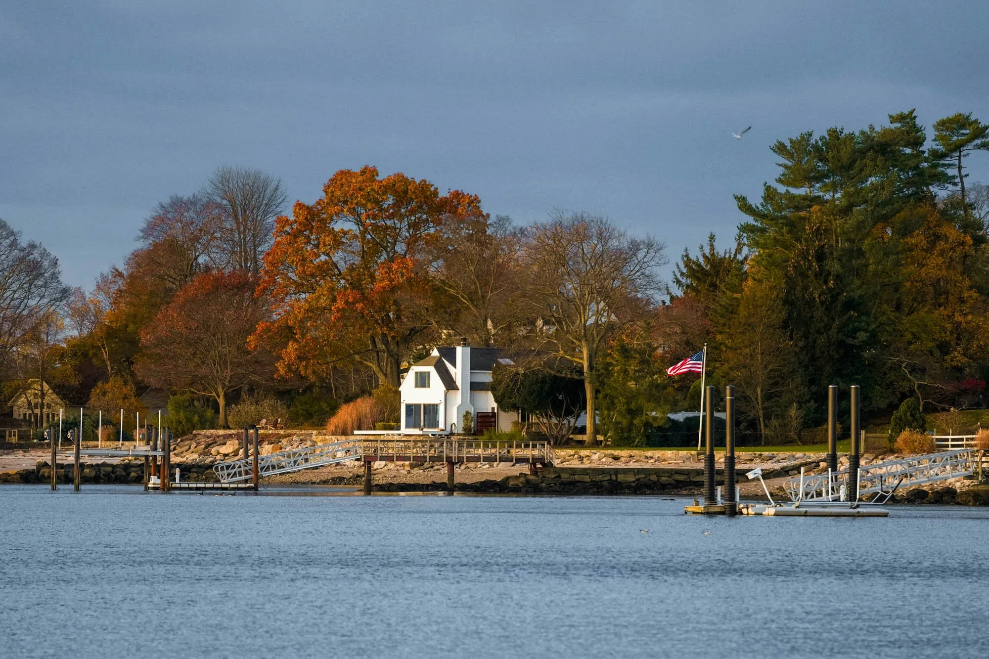 A lakeside scene with a dock and boat ramp, a white house surrounded by colorful trees, and an American flag flying on a pole, with a bird flying overhead.