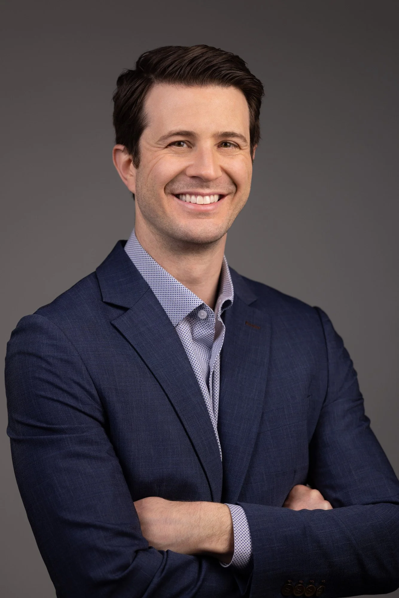 A smiling man with dark brown hair in a professional pose, wearing a navy blue blazer and a light blue dress shirt, standing against a neutral gray background.