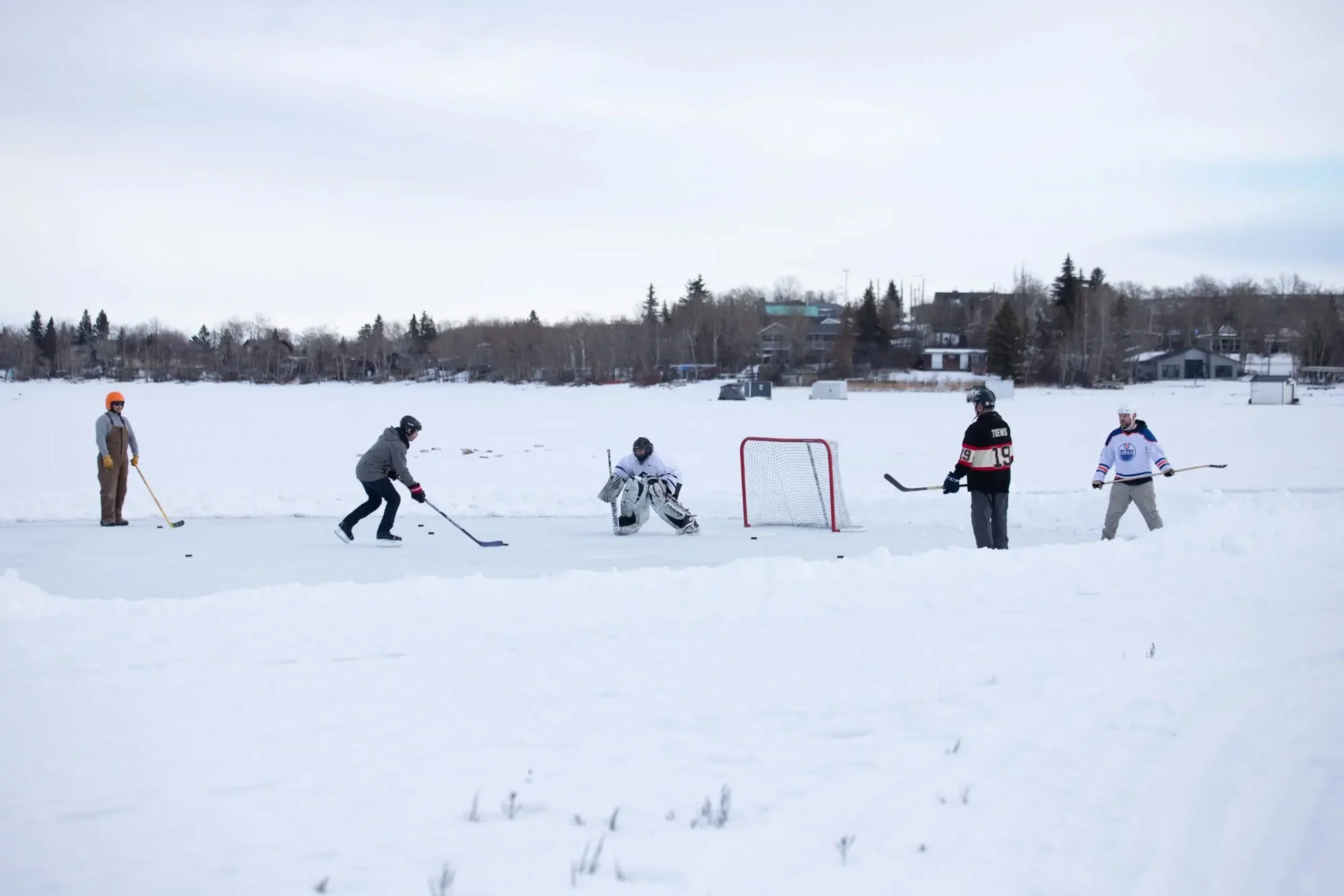 People playing ice hockey outdoors in snowy landscape, with a group around a goal, some with hockey sticks, in a residential area.
