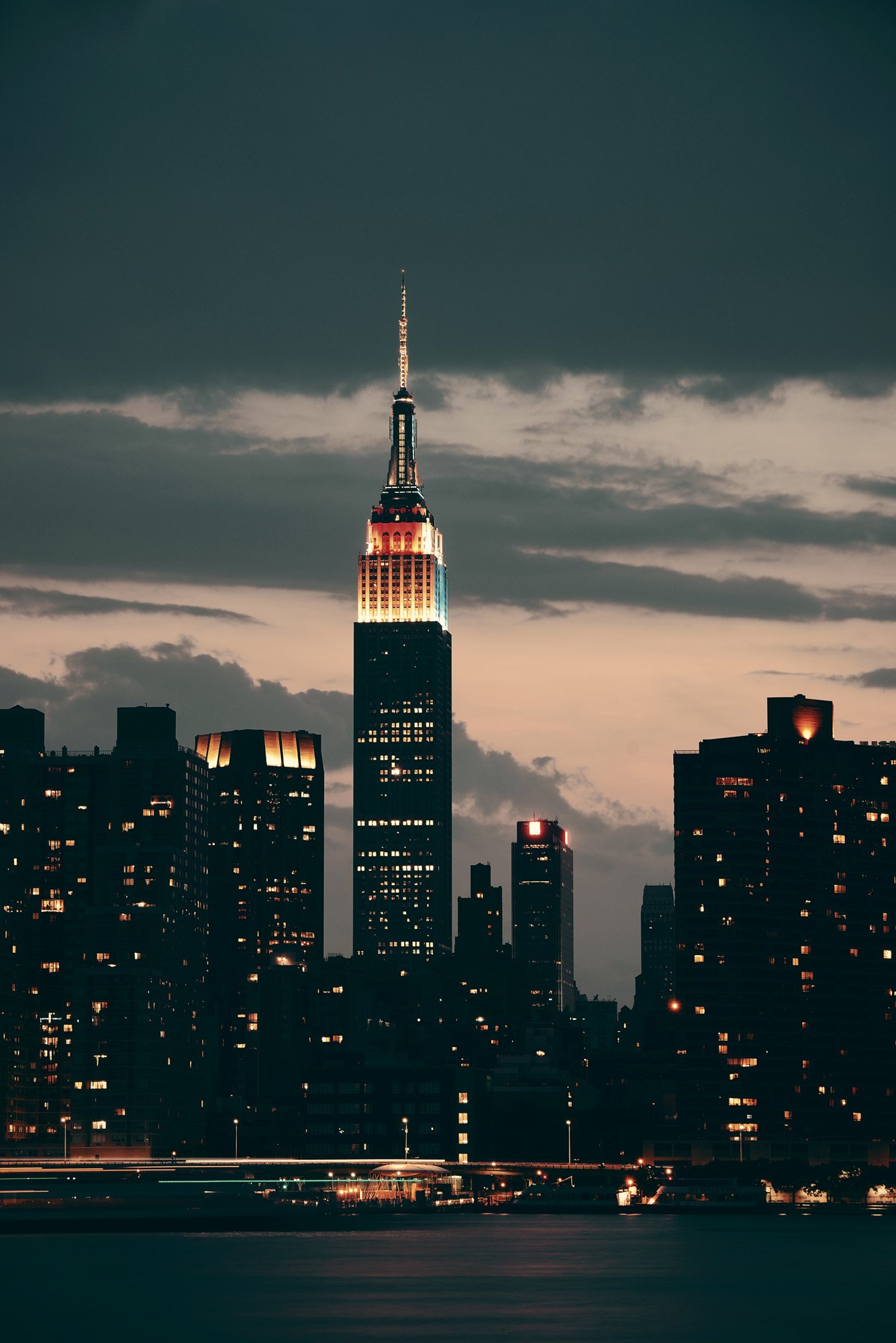 Nighttime cityscape of New York City with the Empire State Building illuminated, surrounded by other tall buildings and dark clouds overhead.