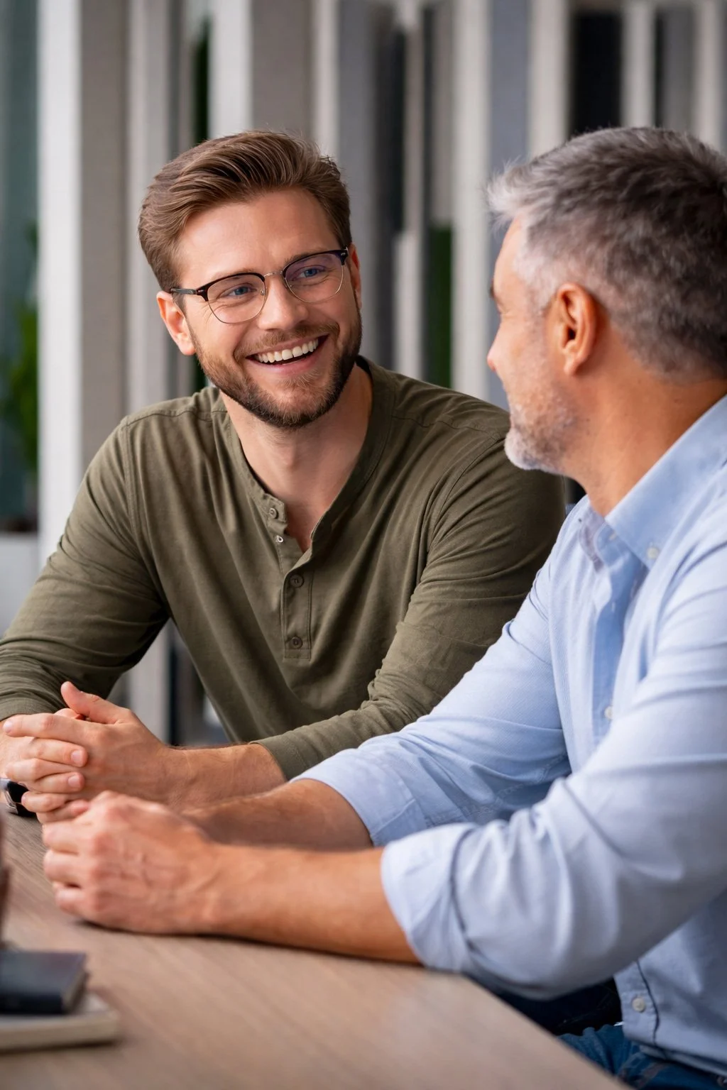 Two men sitting at a table, engaged in conversation and smiling, with one wearing glasses and a green shirt and the other with gray hair and a light blue shirt, in a professional or casual setting.
