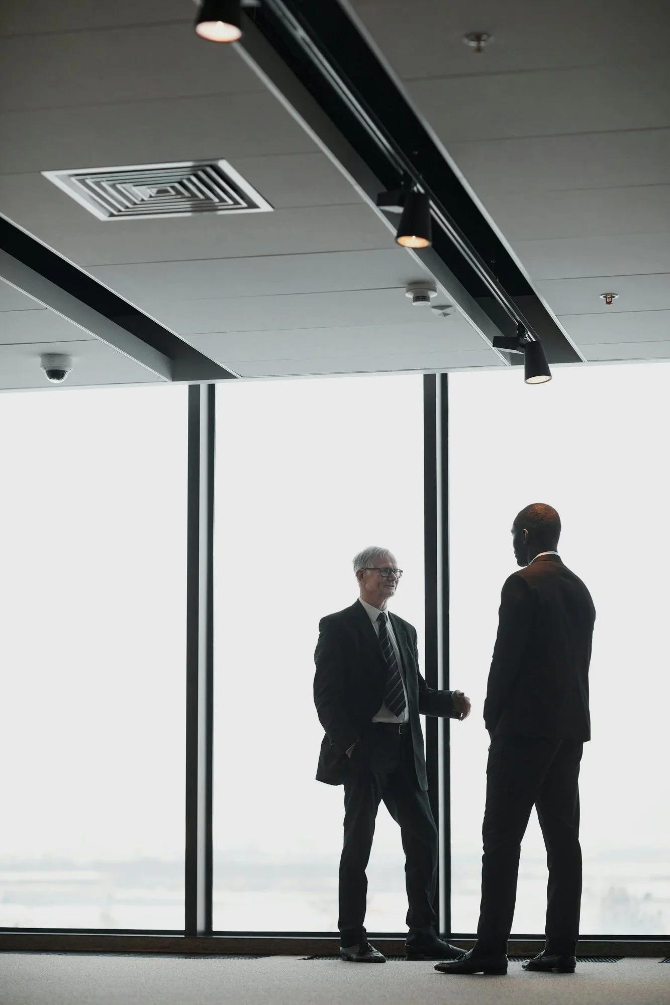 Two business professionals, one with gray hair and glasses and the other Black, are standing and talking in a modern office with large floor-to-ceiling windows. The sky is overcast outside.