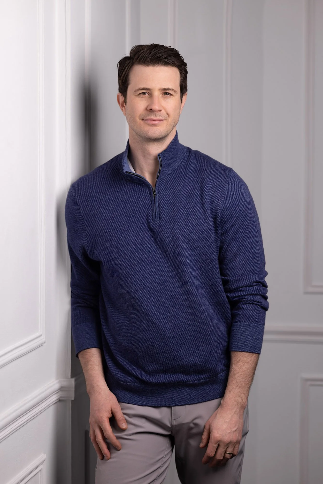 A man with dark hair styled back, standing indoors, leaning against a white wall with decorative paneling. He is wearing a blue quarter-zip sweater and beige pants, looking at the camera with a slight smile.