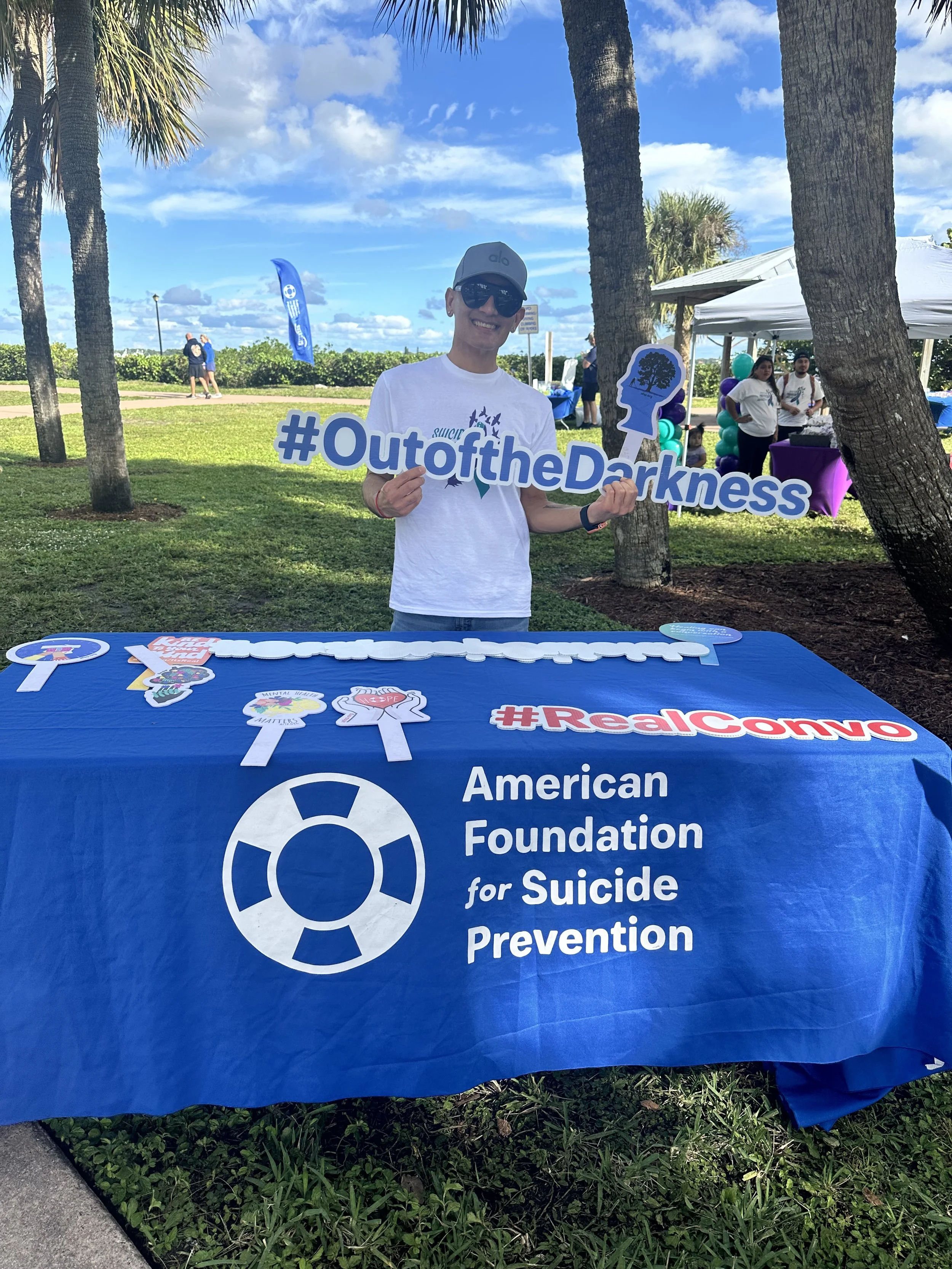 A smiling young man wearing sunglasses and a gray cap holding a sign that reads '#OutoftheDarkness' at a fundraising event for suicide prevention, with a table covered in promotional items, a white tent, and other people in the background under a partly cloudy sky and palm trees.