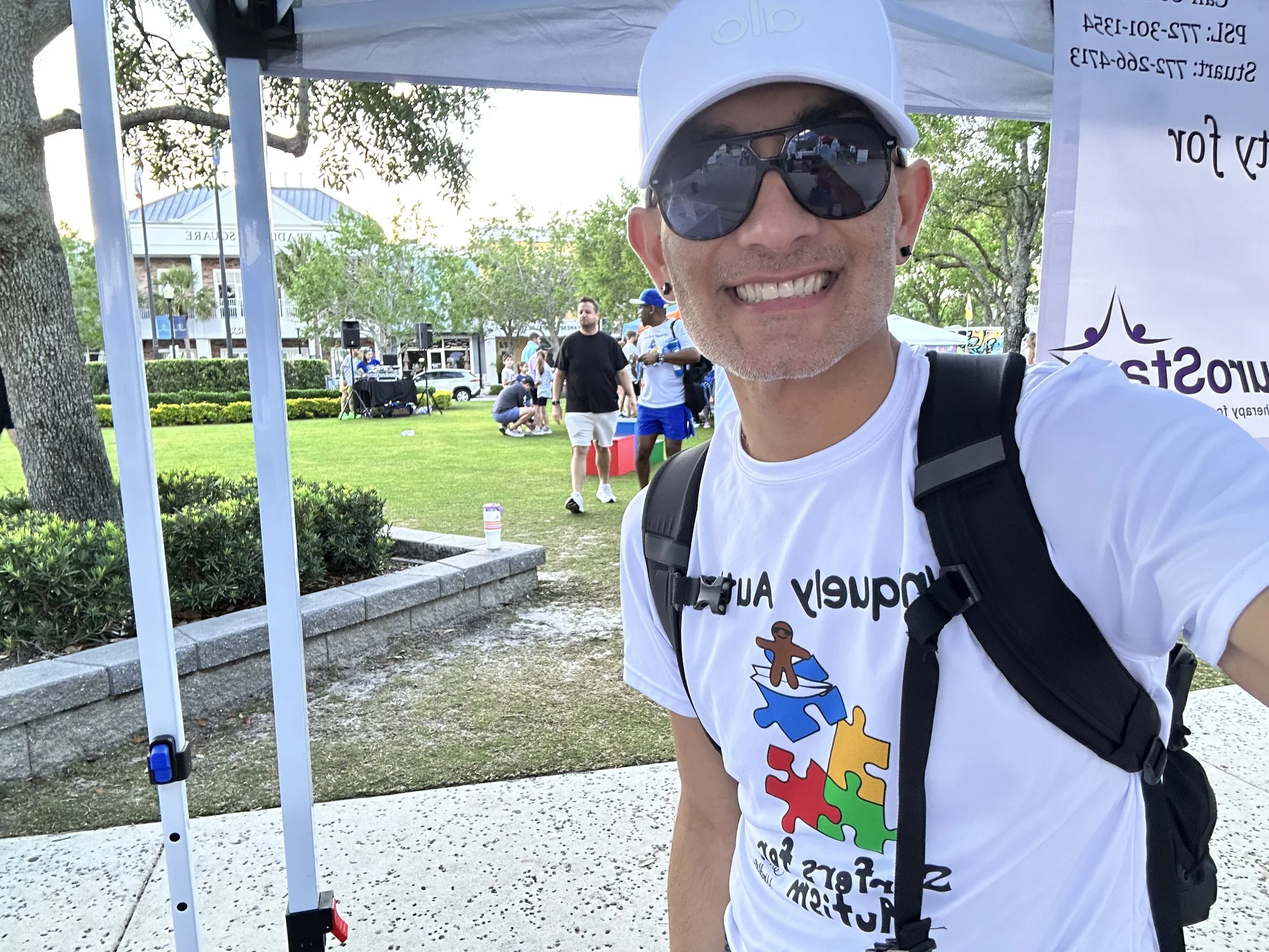 A smiling man wearing sunglasses, a white cap, and a white t-shirt with colorful puzzle piece graphics taking a selfie outdoors at a park event with trees, a building, and people in the background.