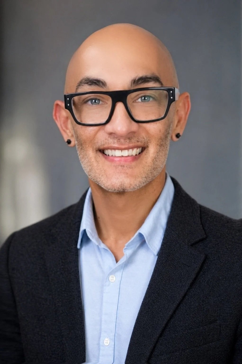 A smiling man with light skin, bald head, glasses, and black earrings, wearing a blue shirt and dark blazer.