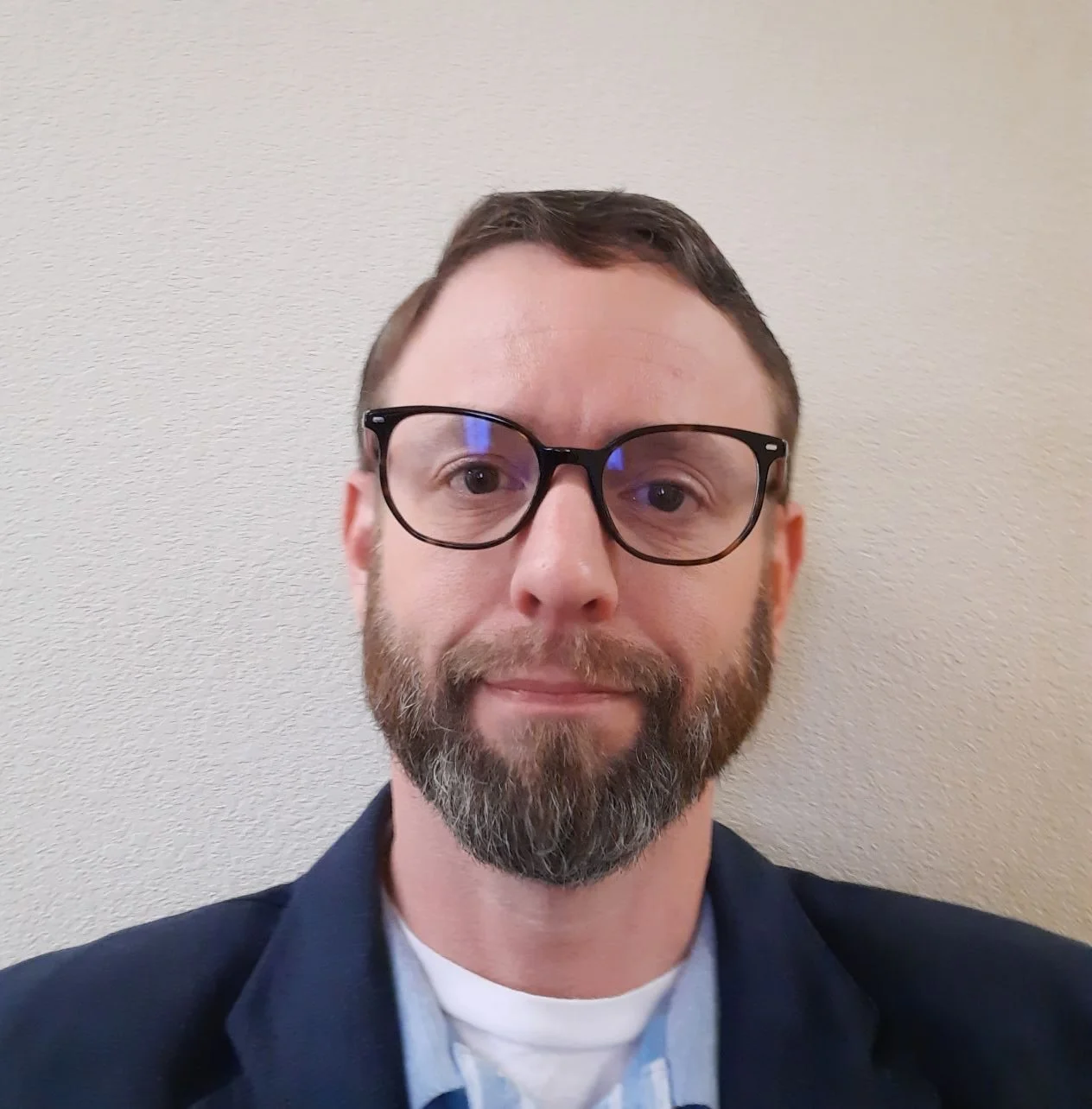 A man with short brown hair, glasses, and a beard taking a selfie against a plain beige wall.