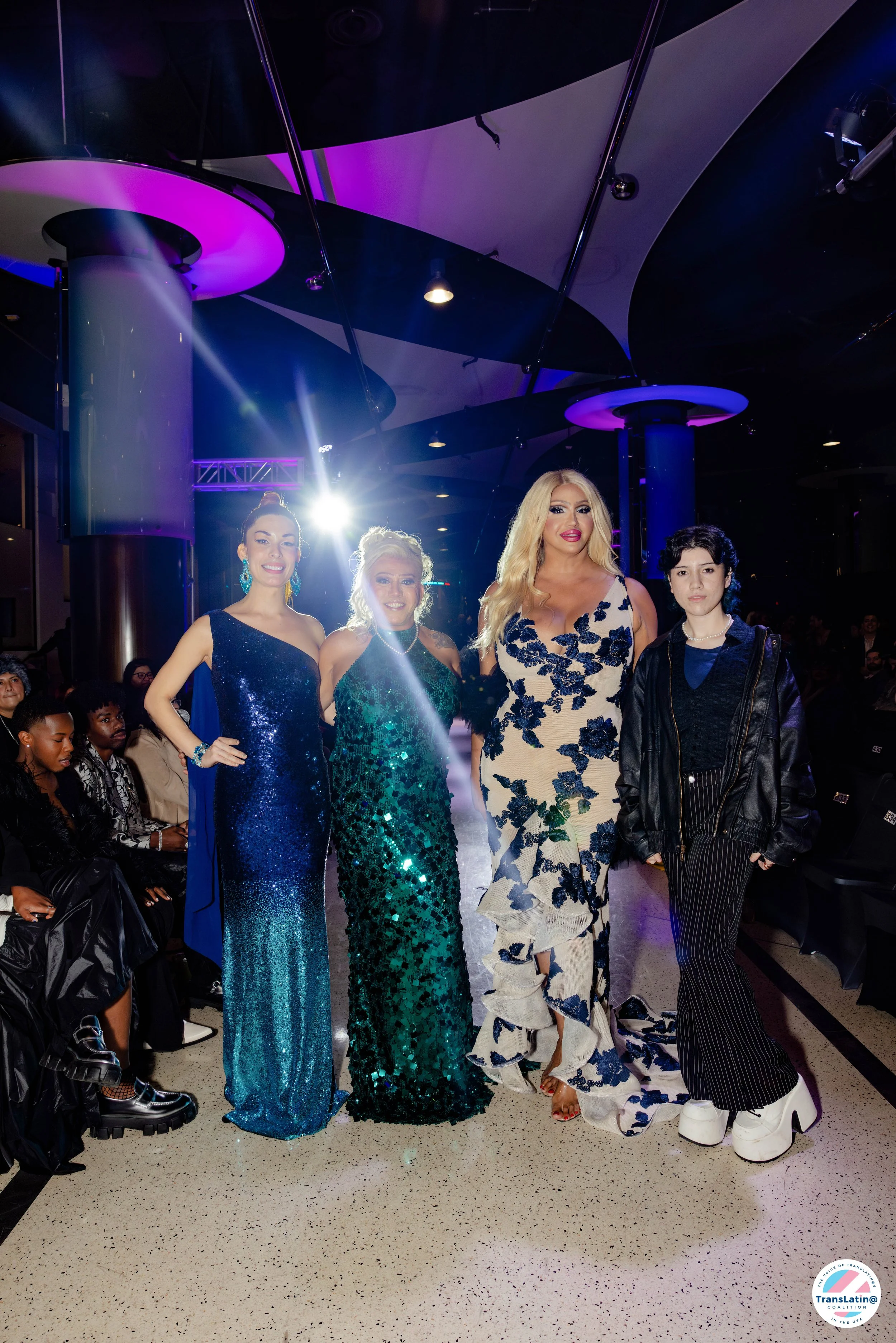 Four women standing together at an indoor event, all dressed in elegant evening gowns, with colorful lighting and audience in the background.