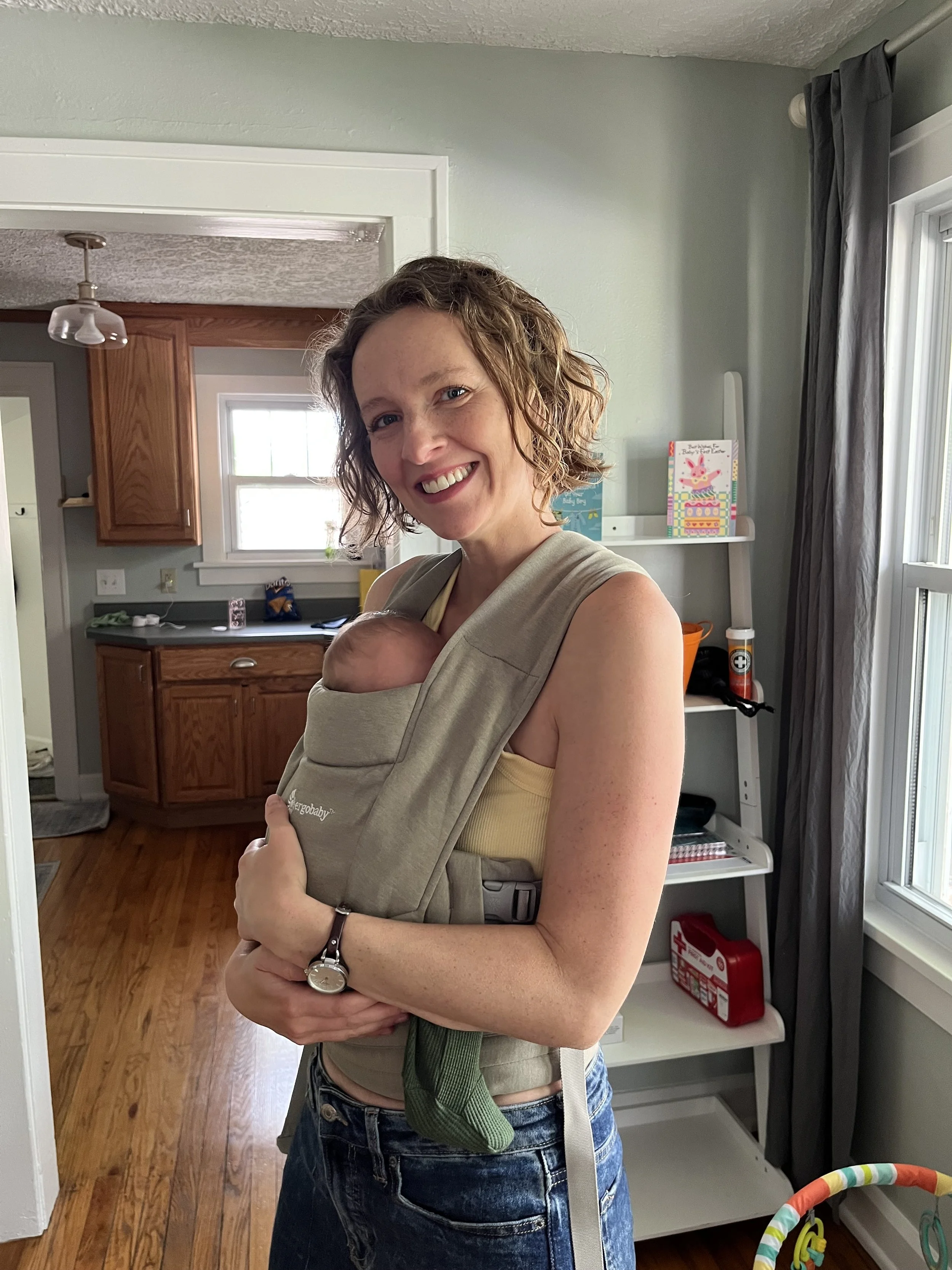 A picture of Grace standing in a home, while wearing a baby in a carrier.