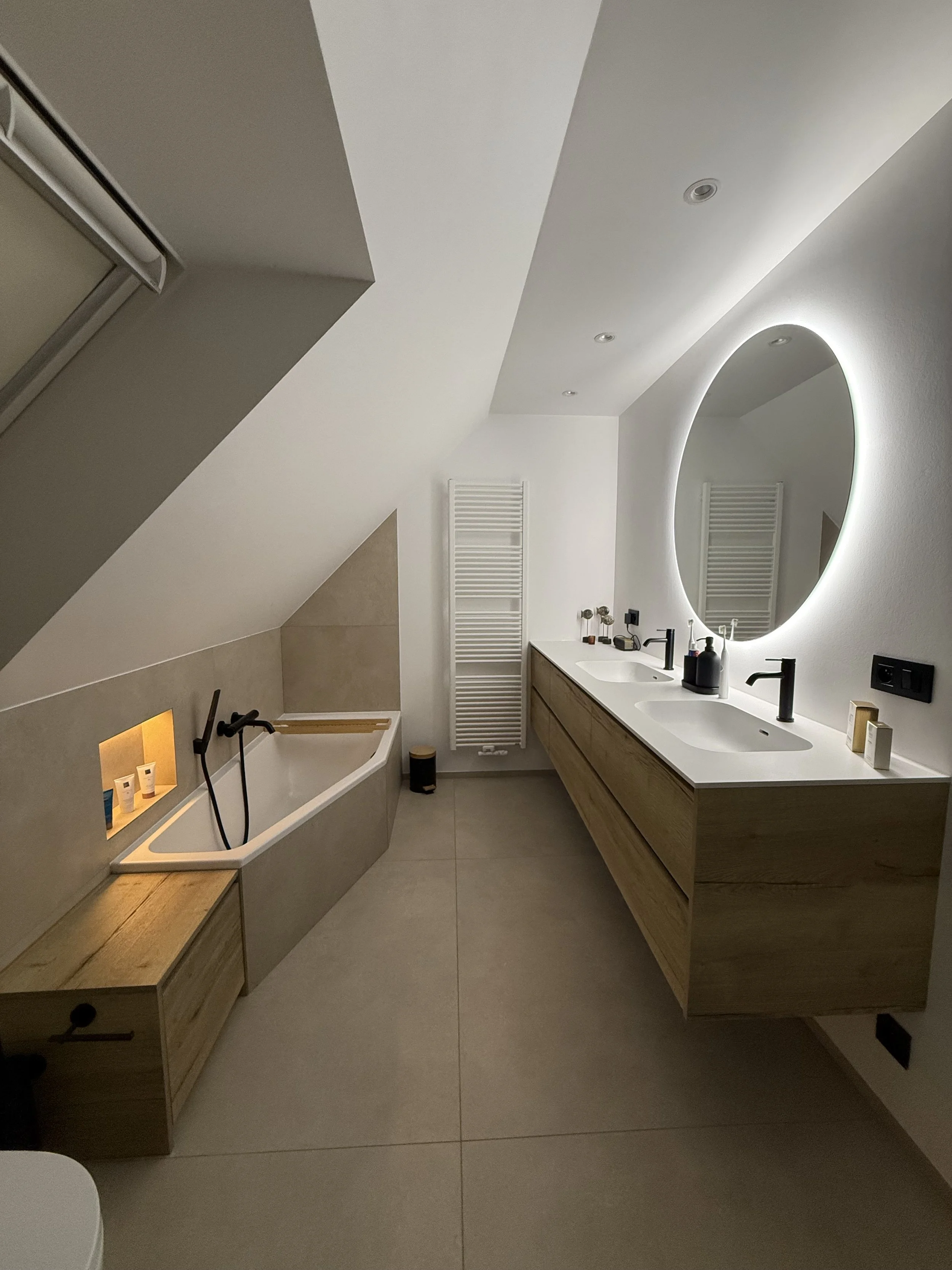 Moderne badkamer met dubbele wasbak, grote ronde spiegel met verlichting, inbouw bad met ingebouwde opbergkast en witte radiator, onder een schuine plafond.