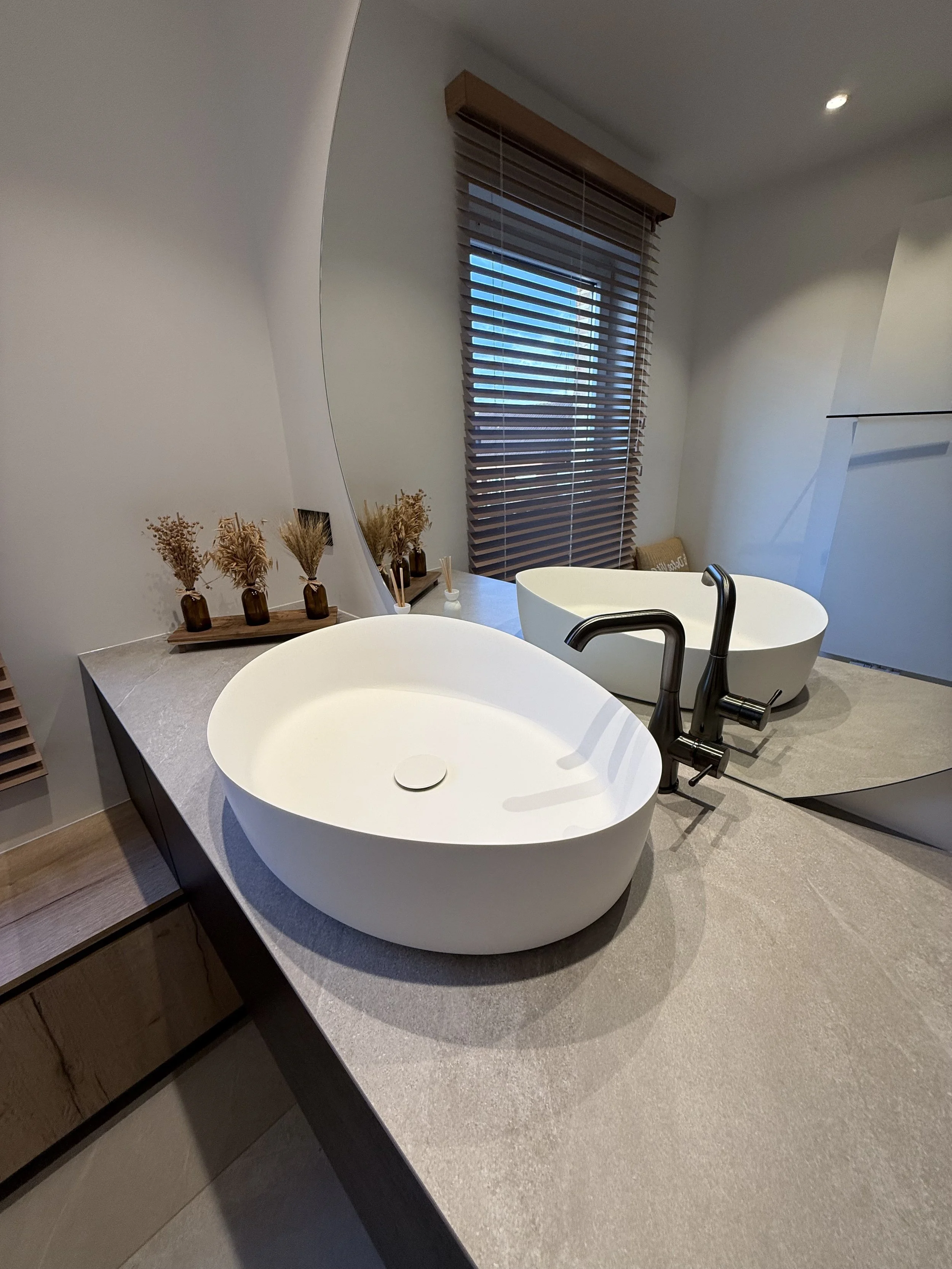 Twee witte vrijstaande wasbekkens met zwarte kranen op een betonlookaanrecht in een badkamer, met een grote spiegel en een raam met houten jaloezieën.