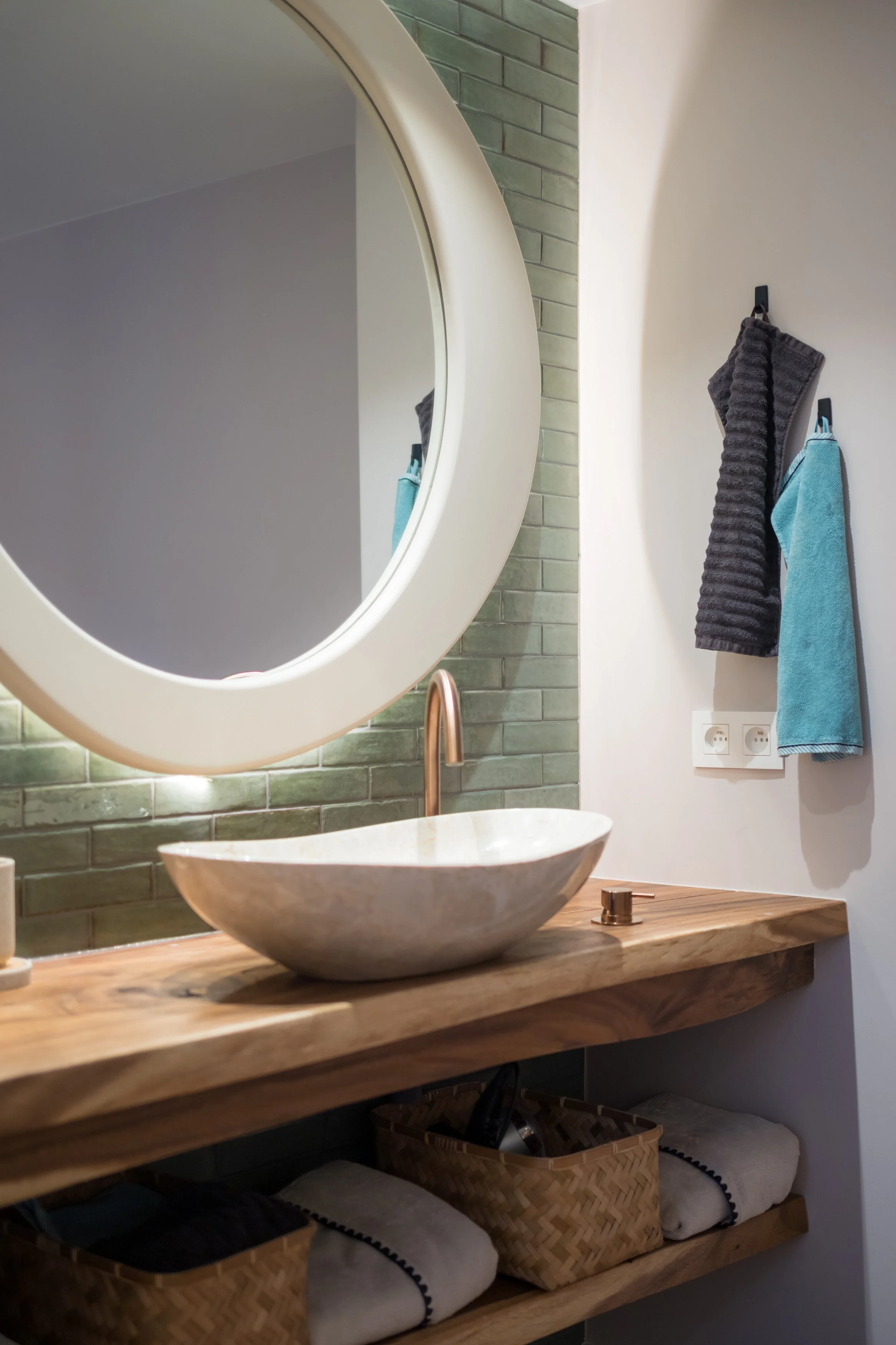 Een badkamer met ruw houten wastafel en een ronde spiegel, groene tegelwand en handdoeken aan de muur.