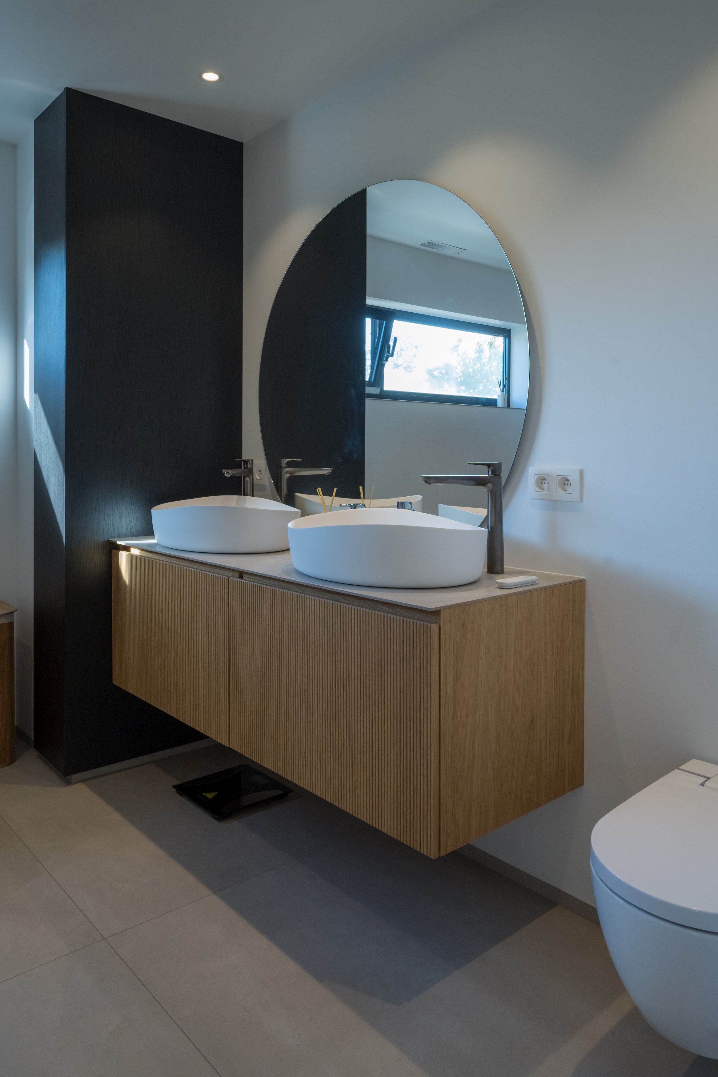 Moderne badkamer met dubbele wasbak, grote spiegel en houten kast onder de wastafels, met een raam voor natuurlijk licht.