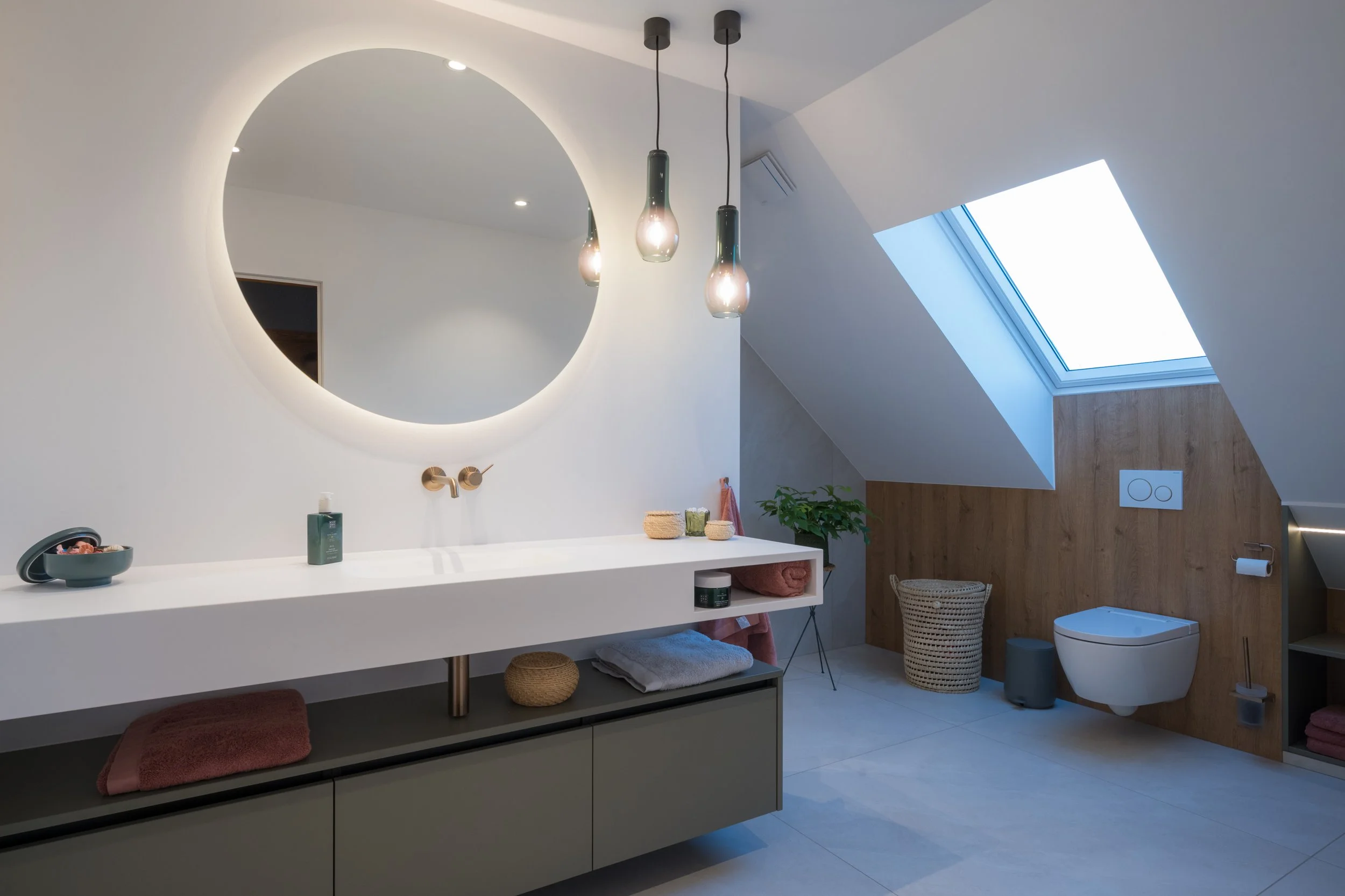 Moderne badkamer met een ronde spiegel, drie hangende lampen, een witte wastafel, een toilet, een skylight in het plafond, en een houten wandachtergrond.