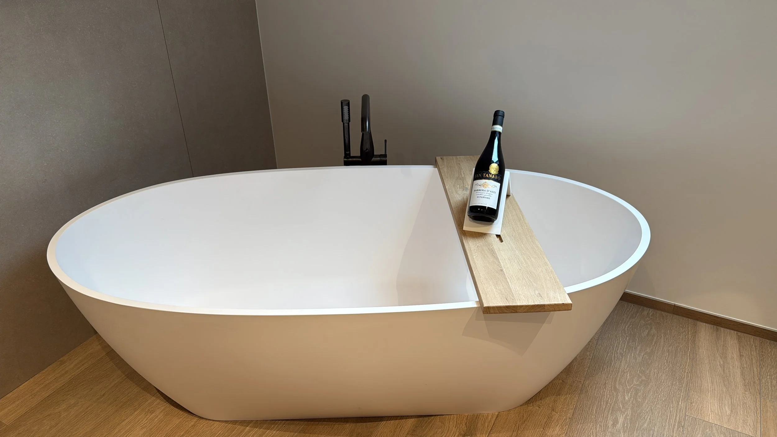 Wit vrijstaand bad op houten vloer met een houten badrand en een fles wijn op een wit bord.