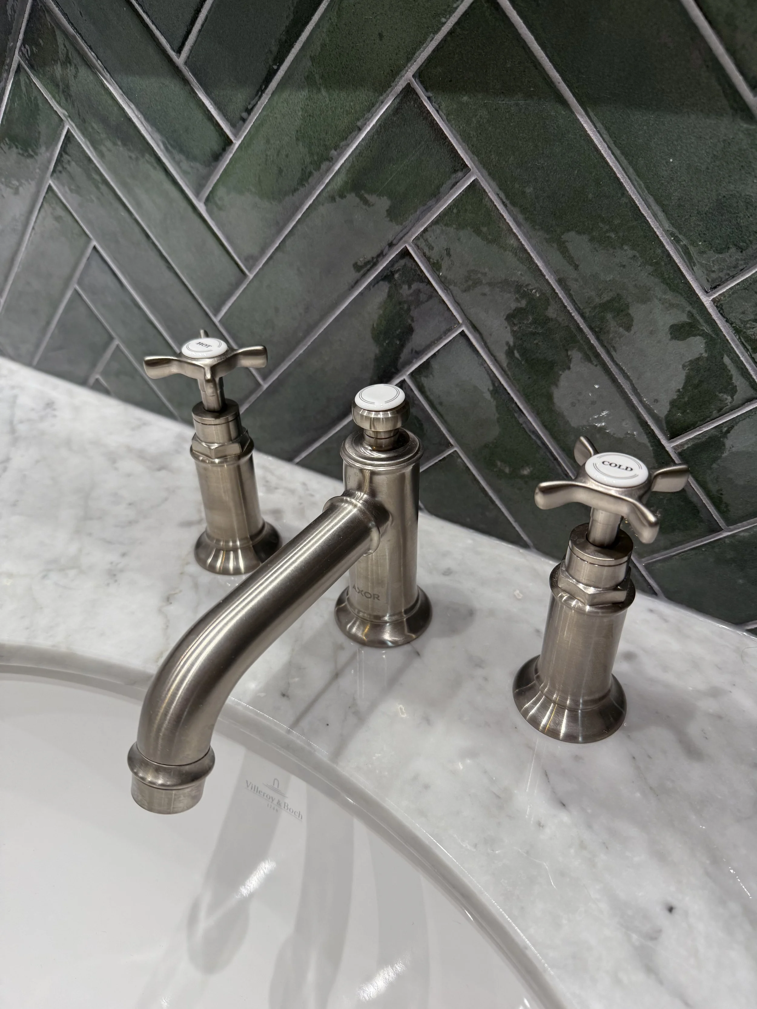 Een badkamer wasbak met een chroomkleurige kraan en twee bijbehorende gemarkeerde handvatten, een met 'COLD' en een met 'HOT', en een marmeren wastafelblad. Op de achtergrond een betegelde muur met donkergroene vierkante tegels.