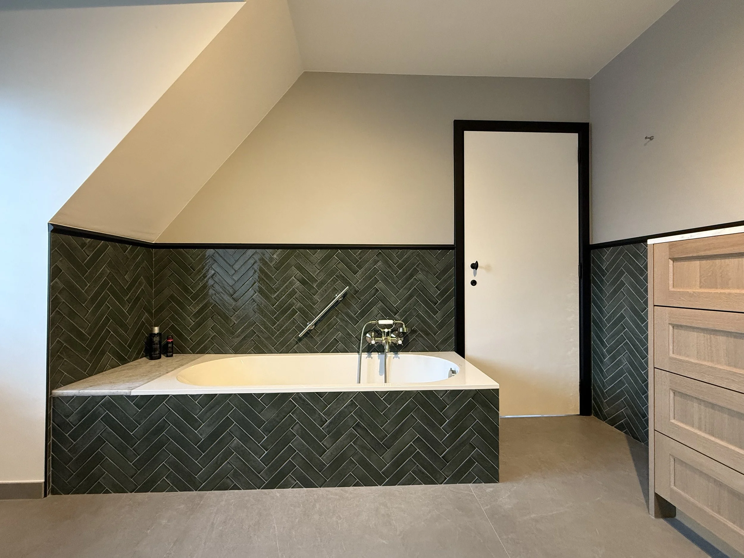 Een badkamer met een ingebouwd ligbad onder een schuine muur, betegeld met donkere visgraatpatroon tegels, een houten ladekast aan de rechterzijde, en een gesloten deur.