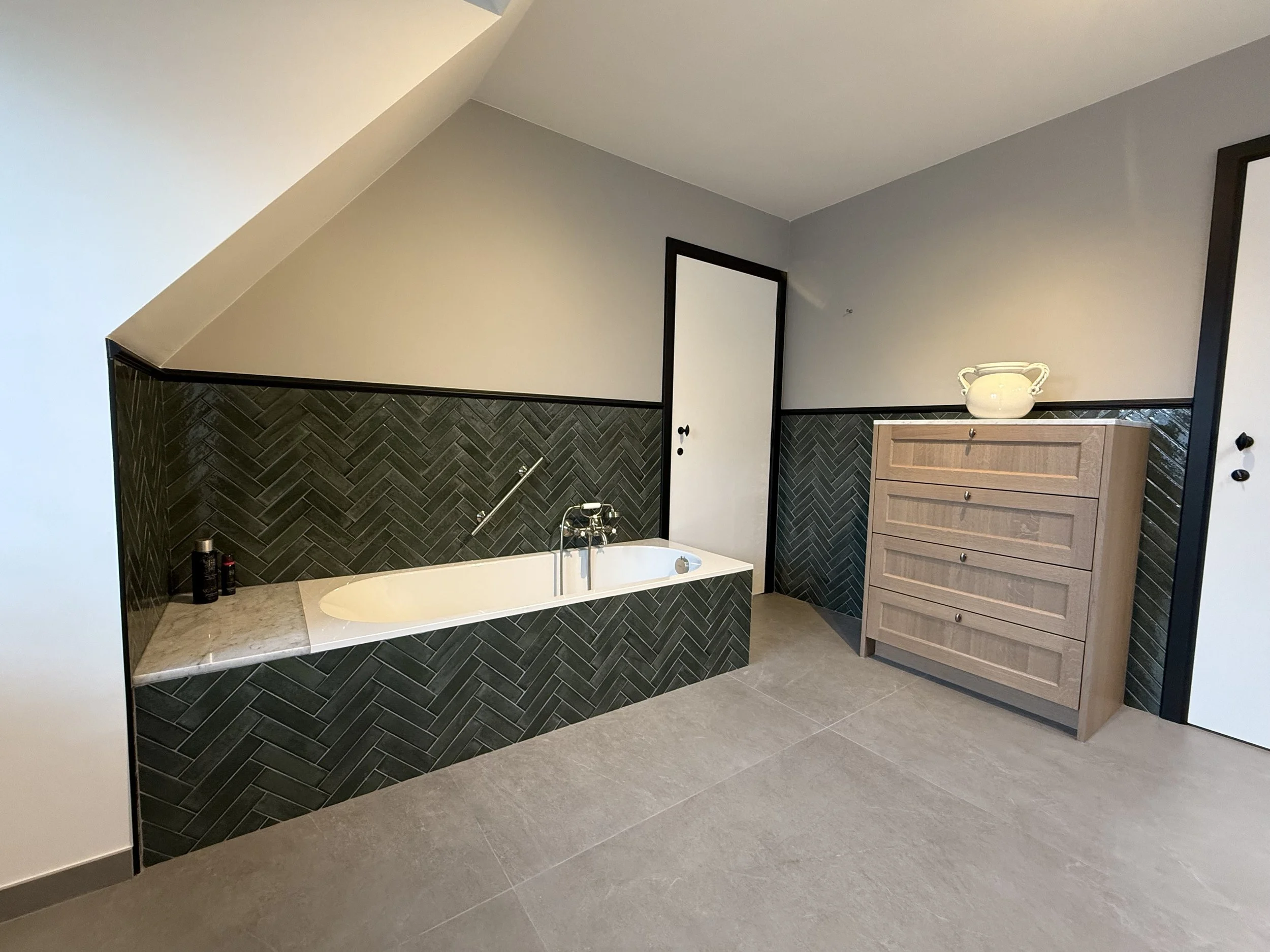 Een inloopbadkamer met een ingebouwd bad onder een schuine muur, zwarte betegeling in een visgraatpatroon, een houten ladekast en een vaas op de kast.