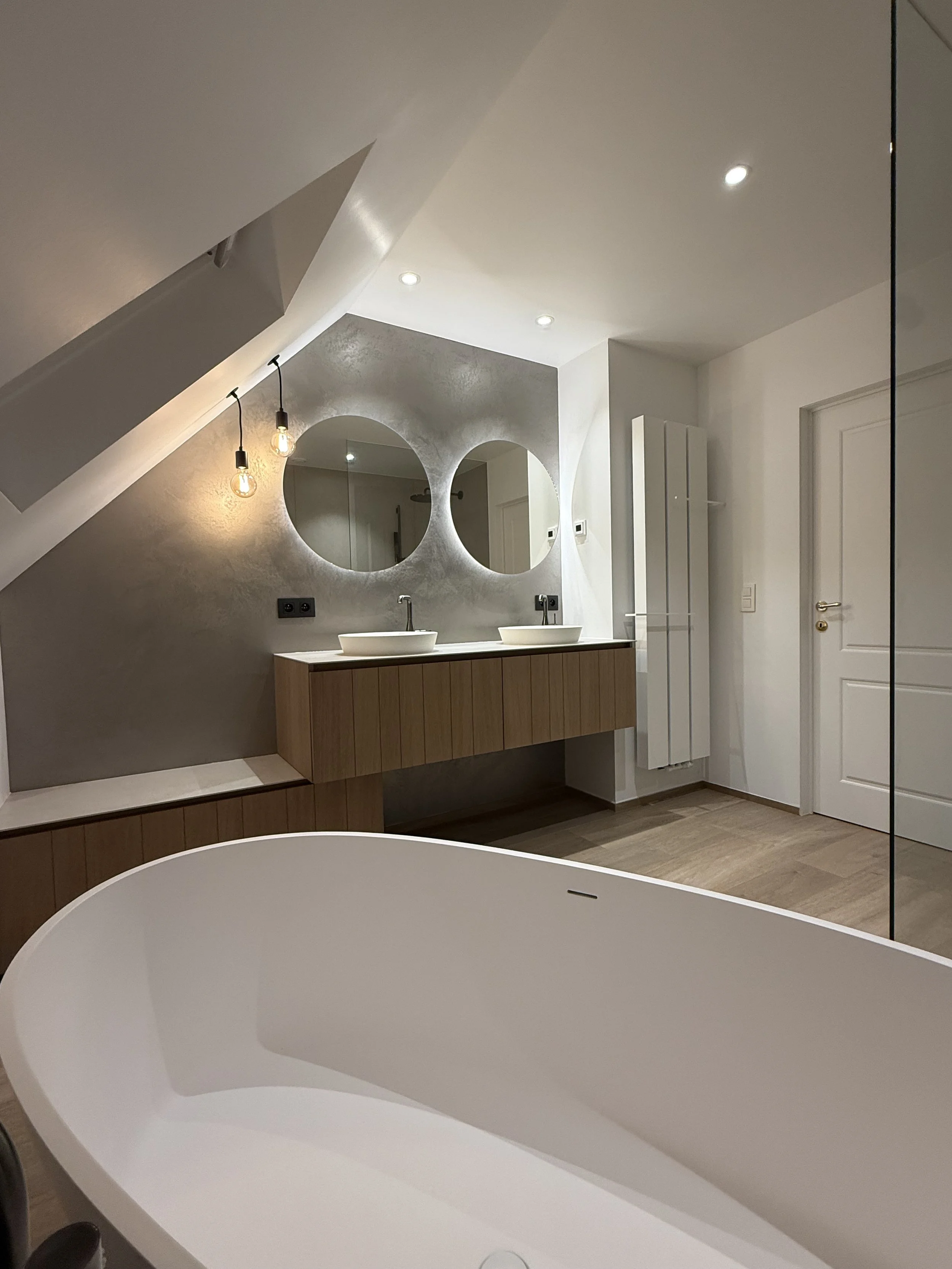 Moderne badkamer met een vrijstaande witte bubbelbad, een houten wastafel met twee waskommen, twee ronde spiegels, plafondverlichting en een wit paneelradiator.