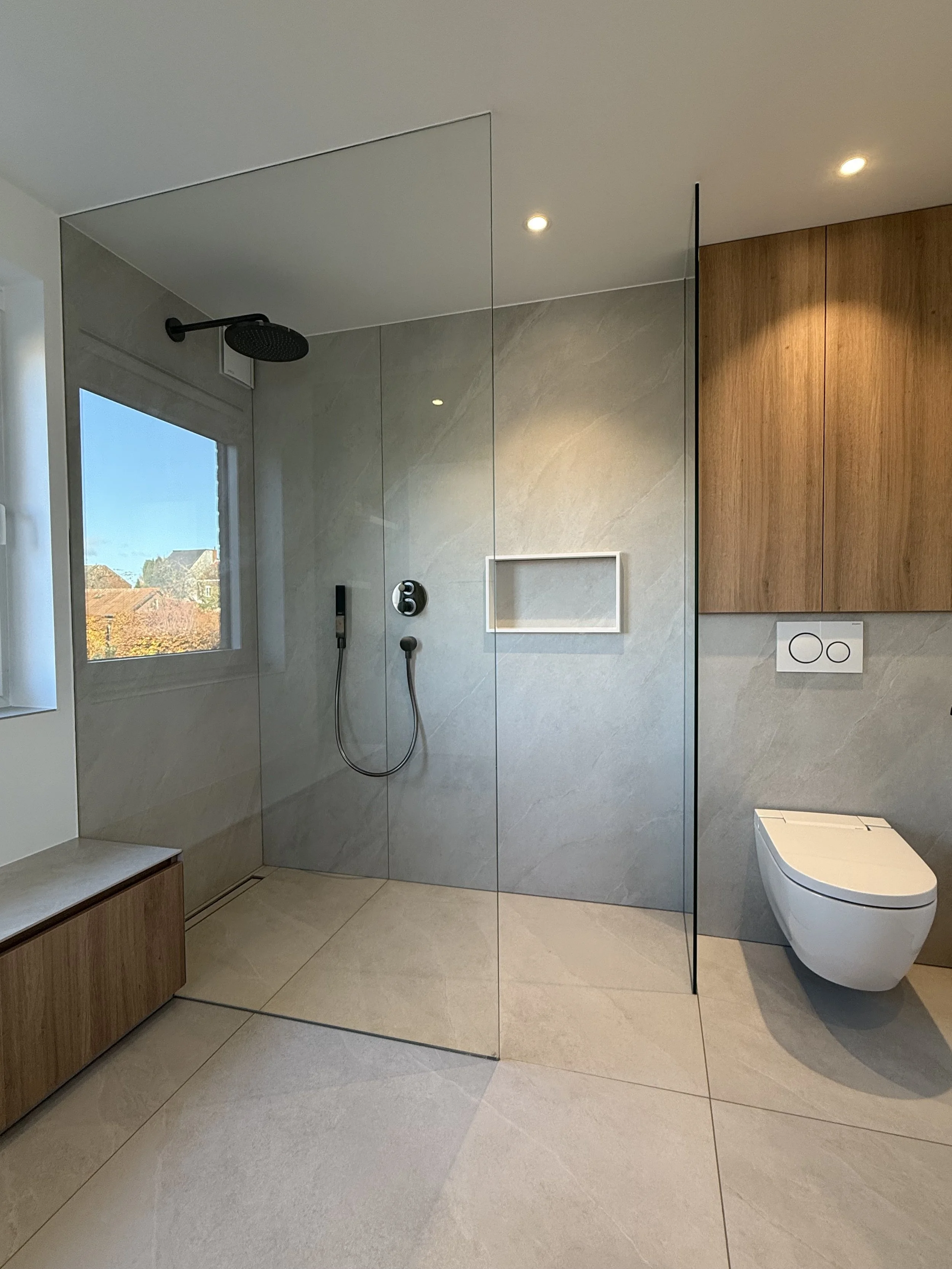 Moderne badkamer met inloopdouche, wandcloset en houten kasten, grote ramen met uitzicht op daken, neutrale kleuren en ingebouwde verlichting.