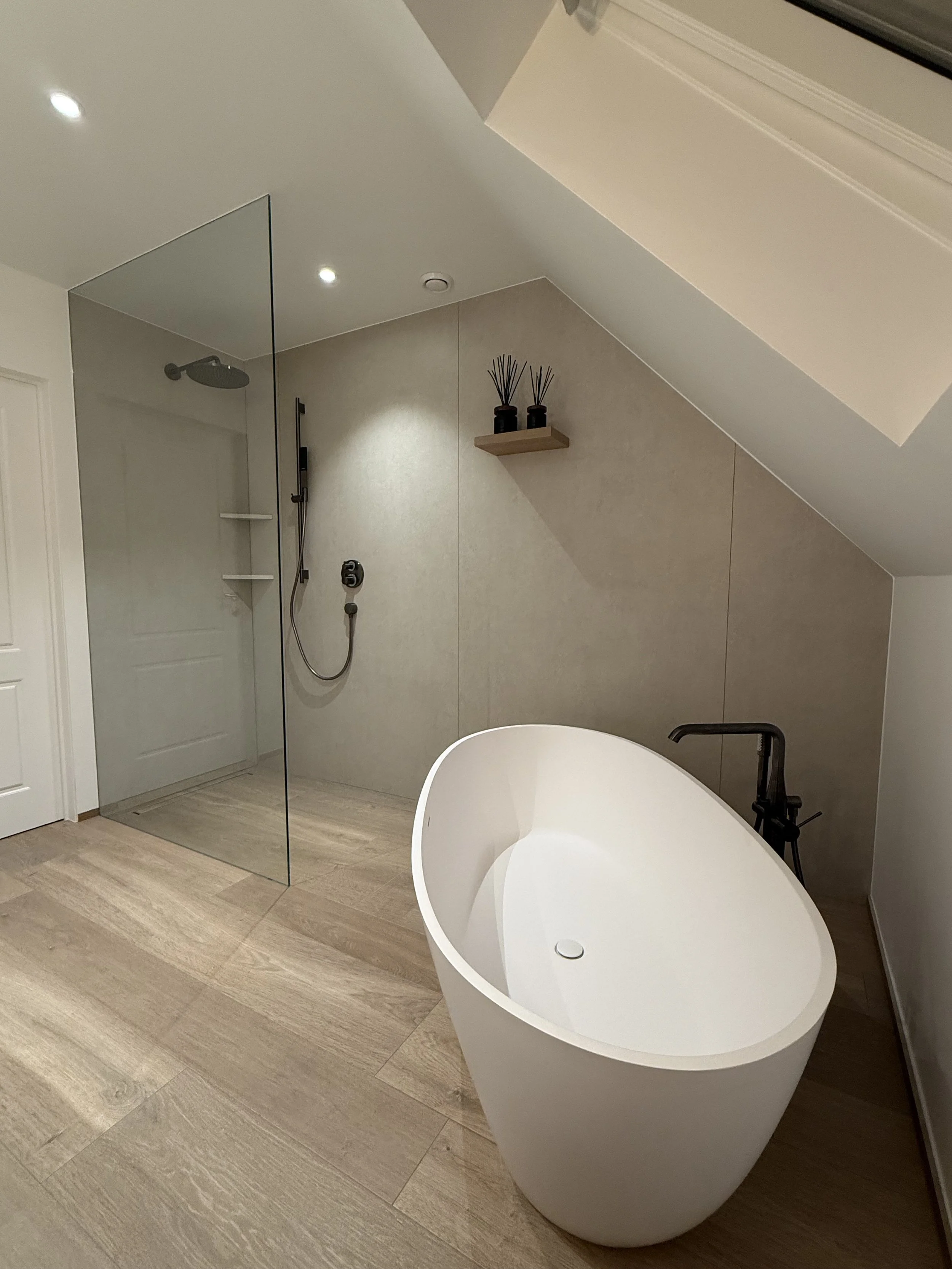 Een moderne badkamer met een vrijstaande badkuip, een inloopdouche met glazen wand, een stuk houten vloer en een kleine witte deur. Er is ook een plafondraam en een gevel met een uitstak van het plafond.