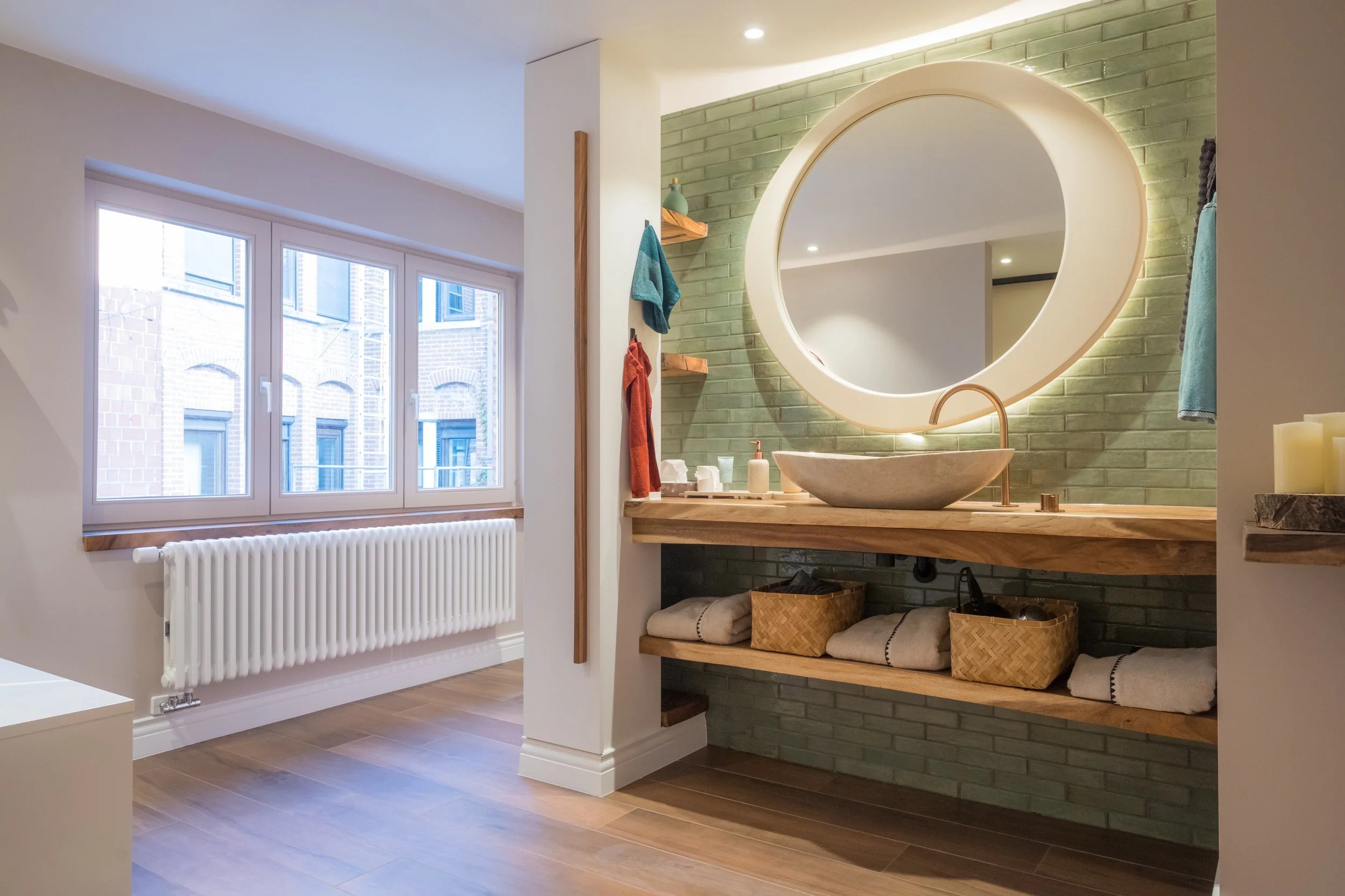 Moderne badkamer met groen bakstenen muur, ronde spiegel met verlichting, houten wastafel met natuurstenen wasbak, handdoeken en schoonmaakproducten, grote ramen met uitzicht op nabijgelegen gebouwen, houten vloer.