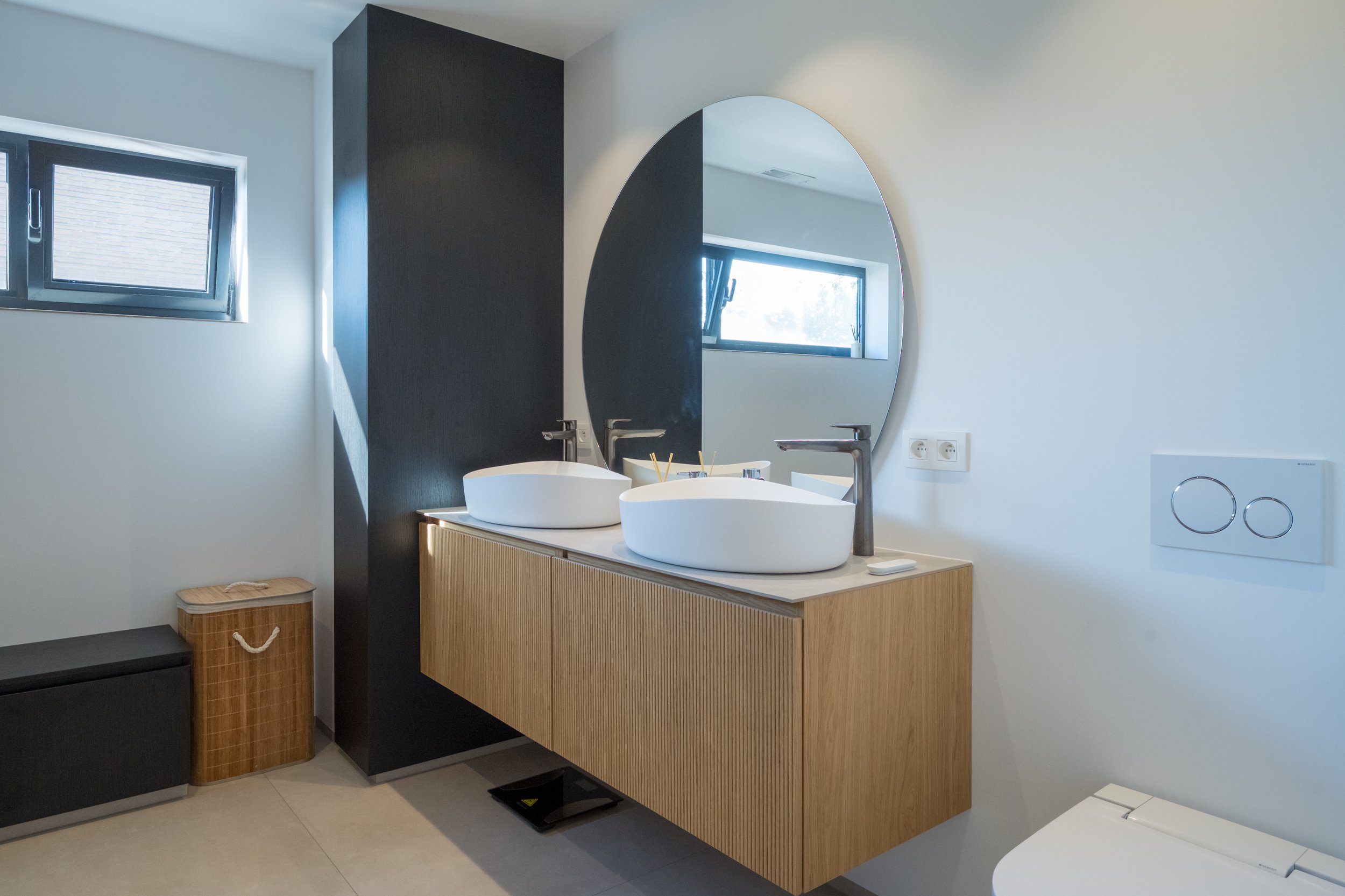 Moderne badkamer met dubbele witte wasbakken, ronde spiegel, zwarte kranen, licht houten kast, kleine venster en beige vloer.