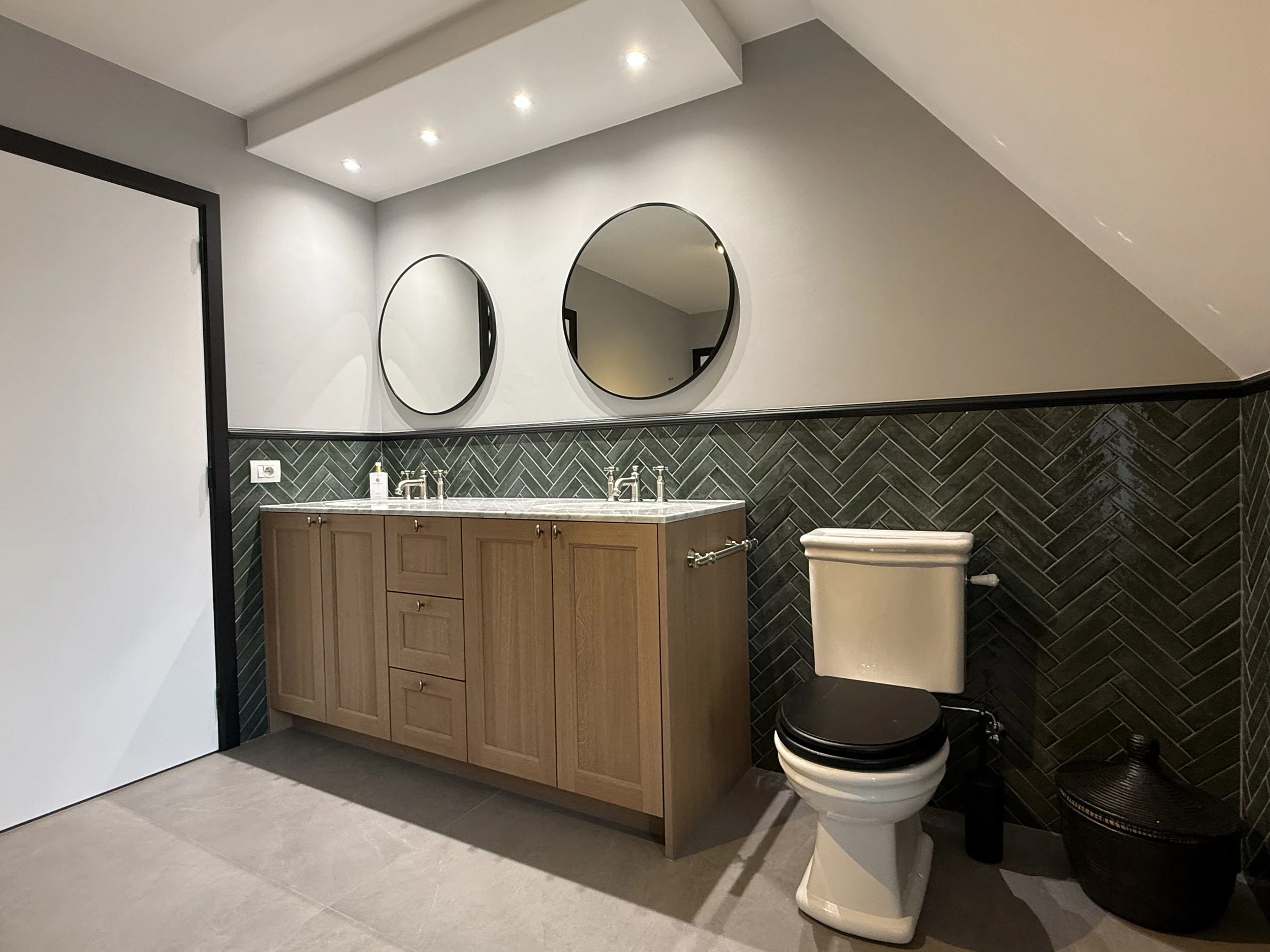 Moderne badkamer met dubbele spiegel, houten kast, toilet en witte muren met zwarte en groene tegelwand.