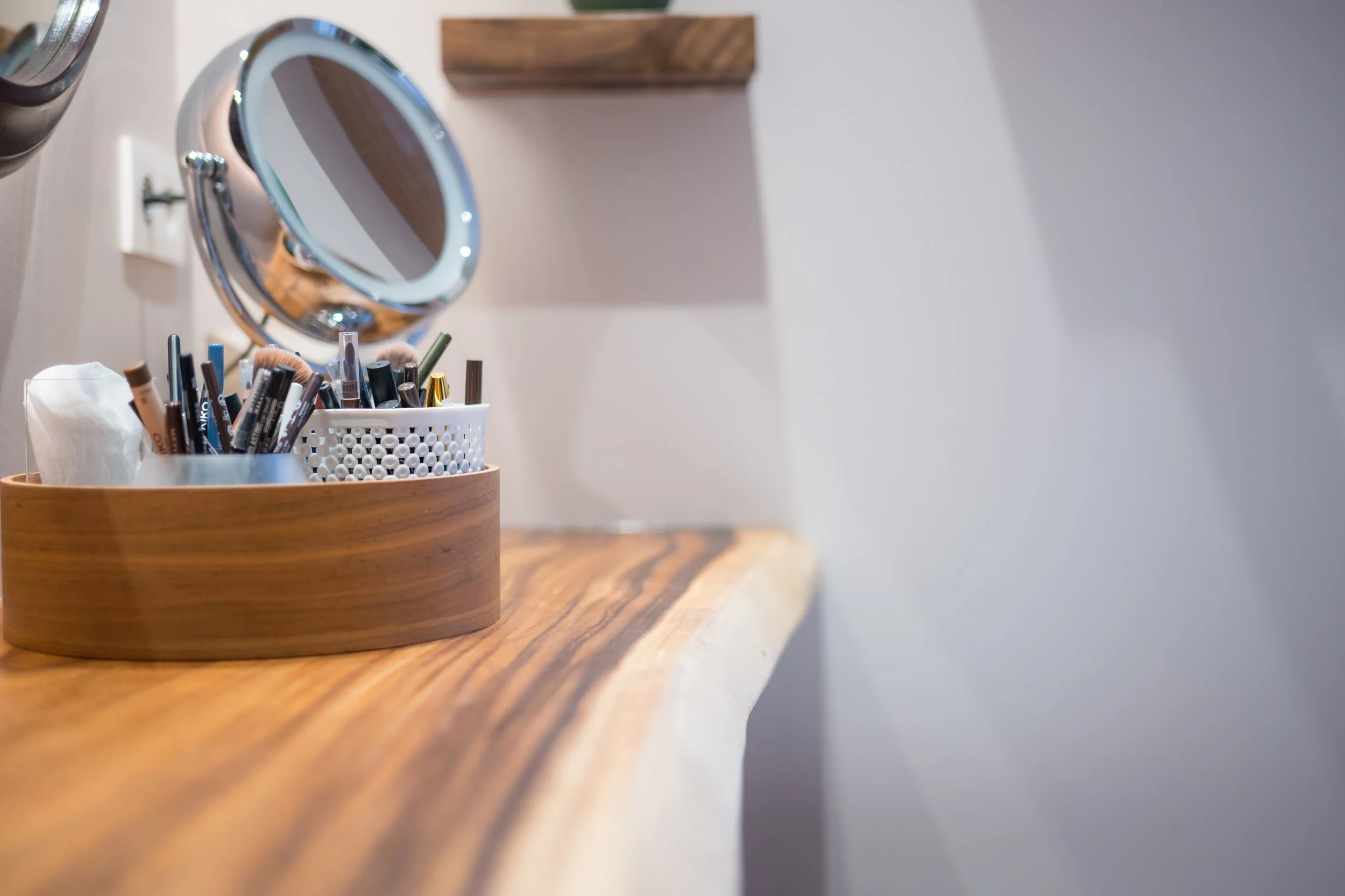 Een houten tafel met een ronde container gevuld met make-up kwasten en oogpotlood. Op de muur hangt een ronde spiegel.