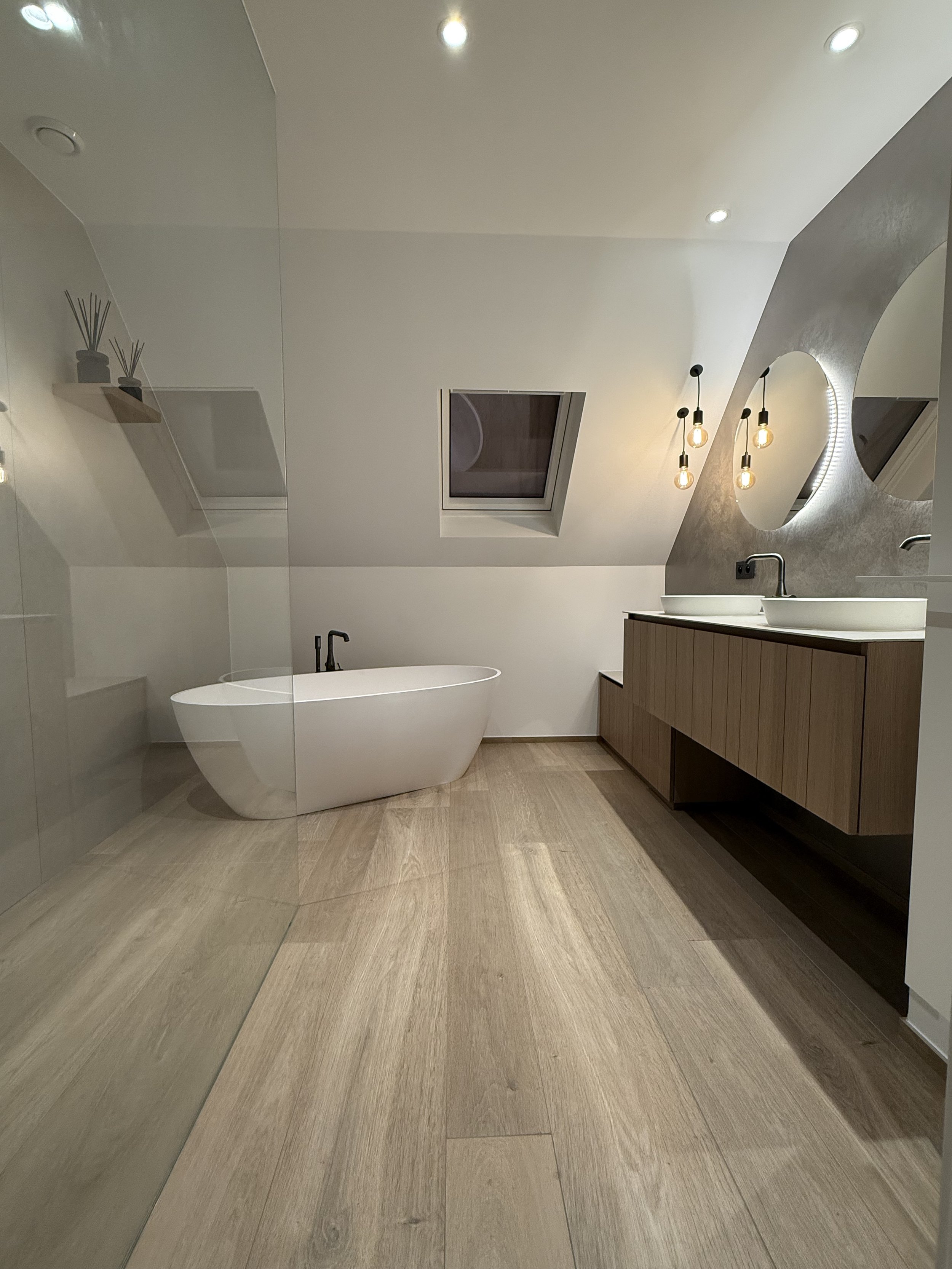 Moderne badkamer met vrijstaande douche en bad, dubbele wastafel met ronde spiegels, houten kast, lichtgekleurde houten vloer, plafond met ingebouwde verlichting en een dakraam.