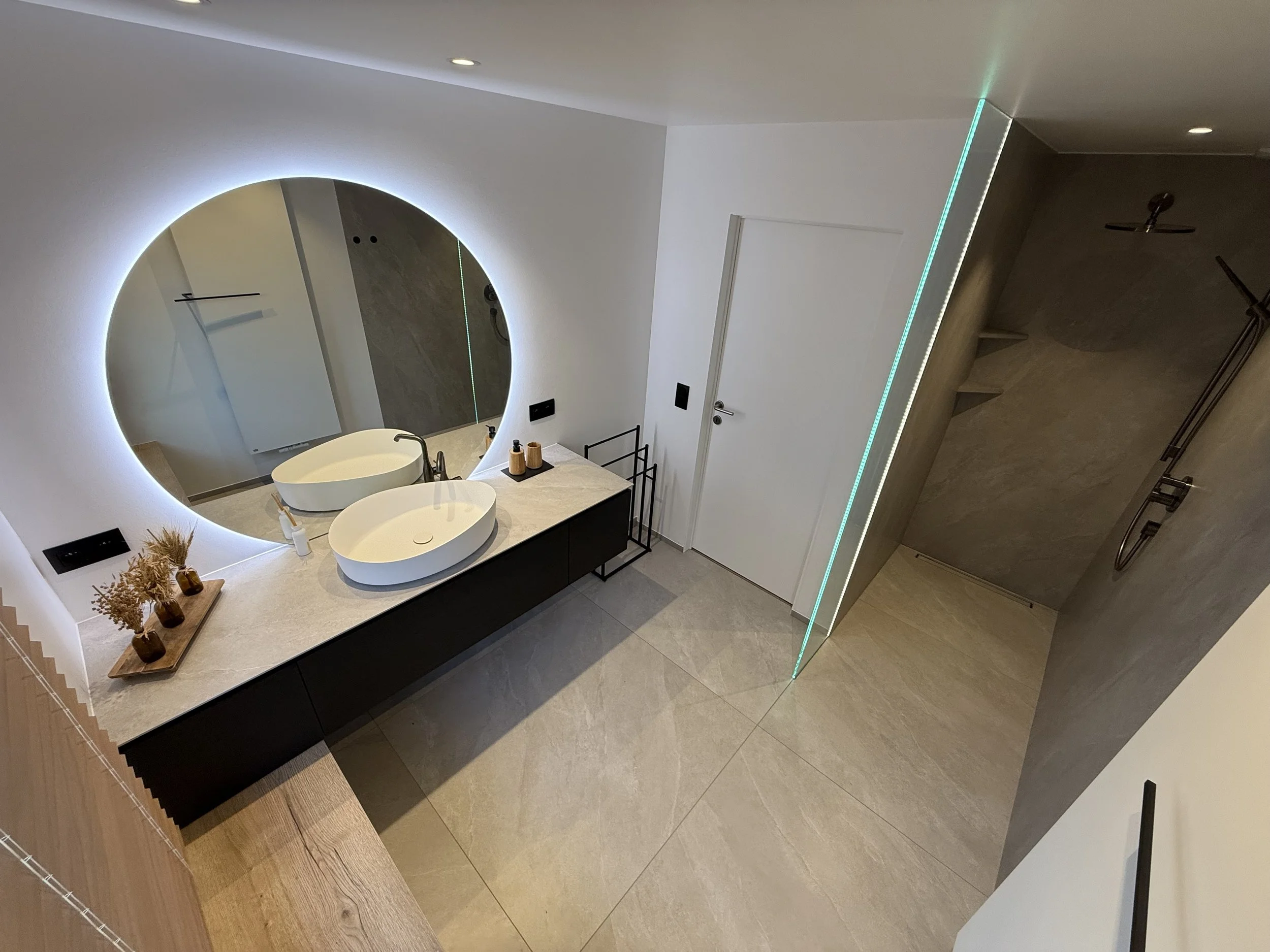 Moderne badruimte met dubbele wit wasbakken, grote ronde spiegel met led-verlichting, een doucheruimte met een regen douche en beige tegels.