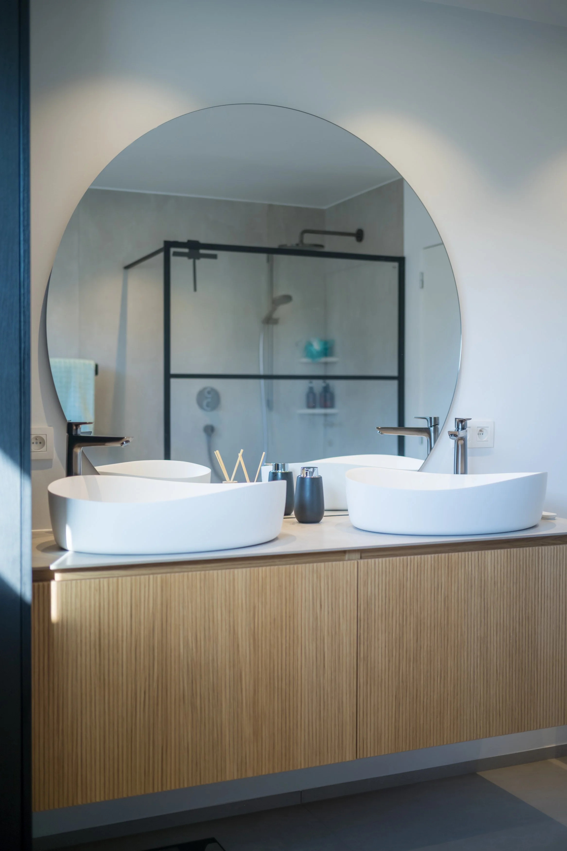 Een badkamer met twee witte wastafels op een houten kast, een grote ronde spiegel, en een inloopdouche in de achtergrond.