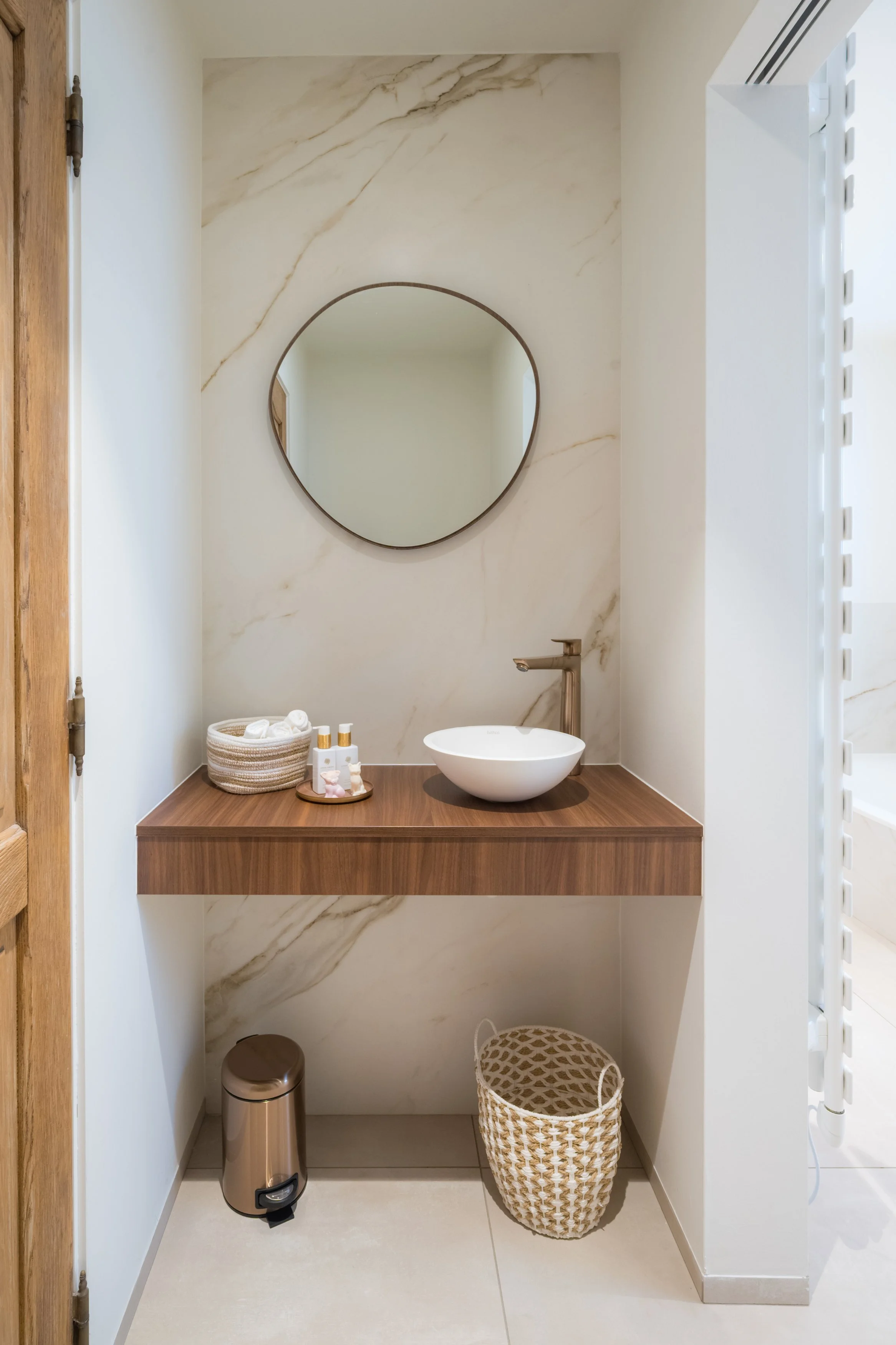 Een badkamer met een ronde spiegel, een witte kommetjes, handdoeken, verzorgingsproducten, een wastafel en een kleine prullenbak.