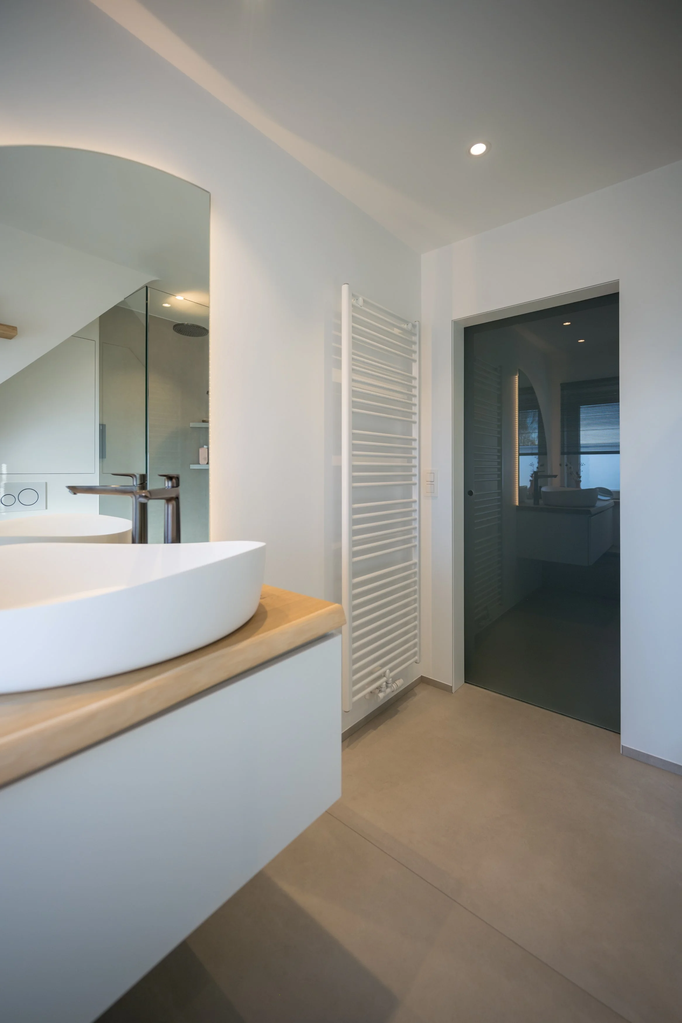Moderne badkamer met witte muur, oven, wit wastafel met houten blad, spiegel, radiator, deur naar andere kamer, en inloopdouche met glazen wand.