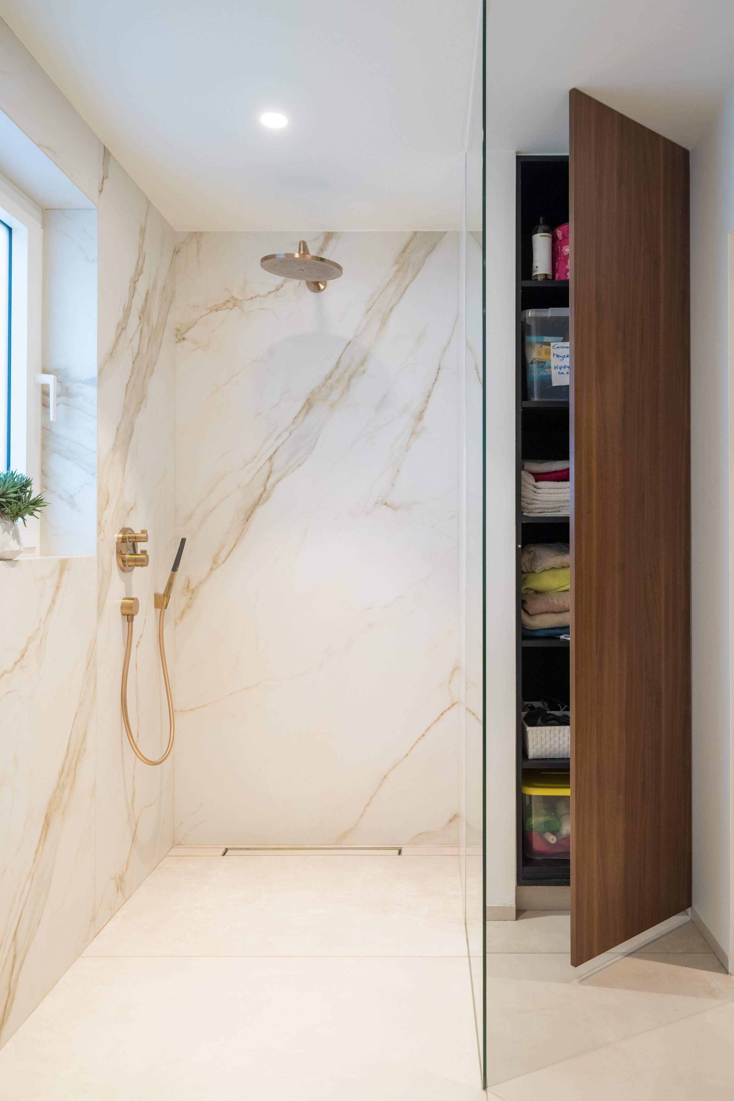 Een moderne inloopdouche met marmeren wandtegels, een regen(douchekop, en een inbouwkast met opgerolde handdoeken en toilettes.