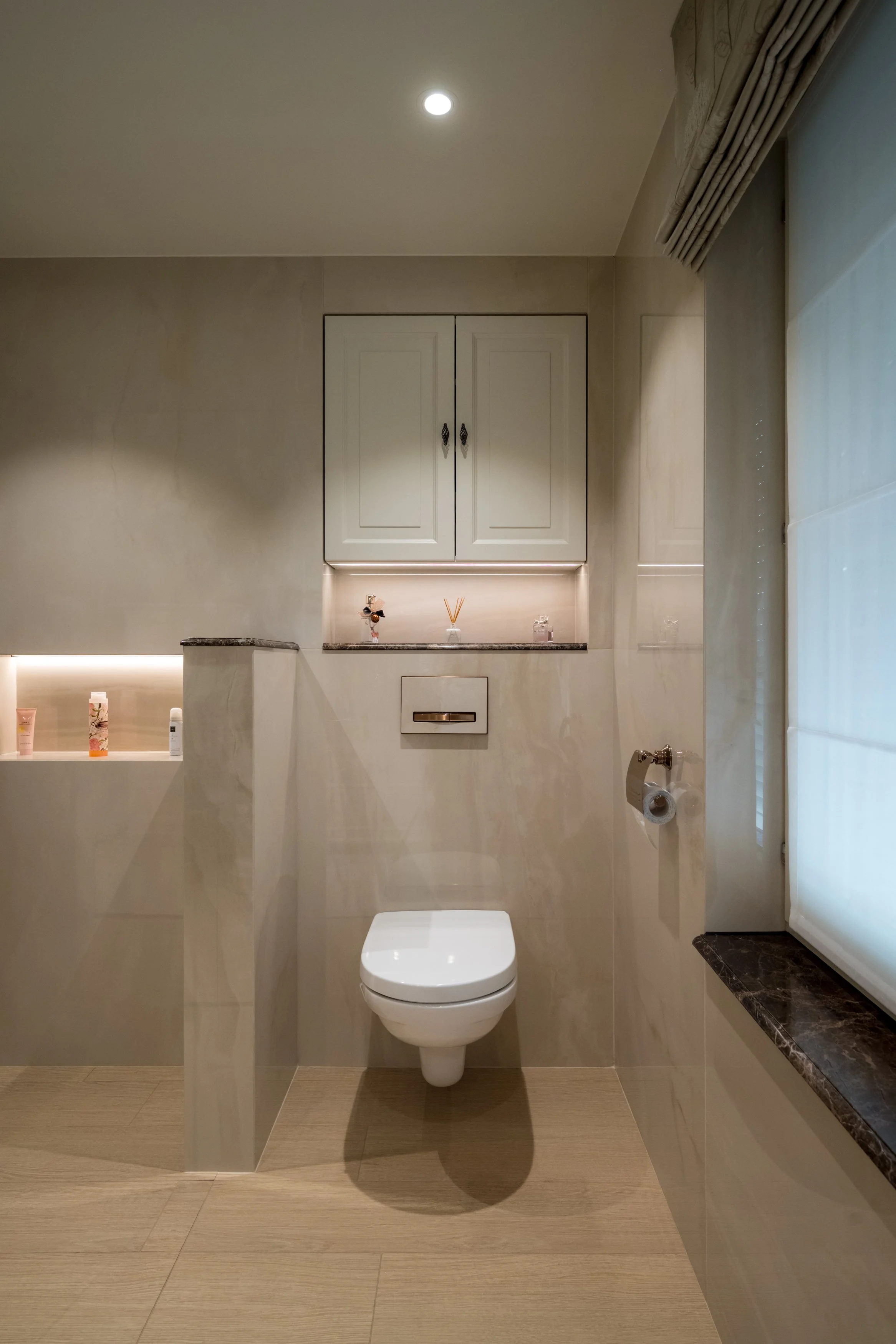 Een moderne, minimalistische badkamer met een wandtoilet, een wit kastje boven het toilet, en een raam met een gordijn aan de rechterkant.