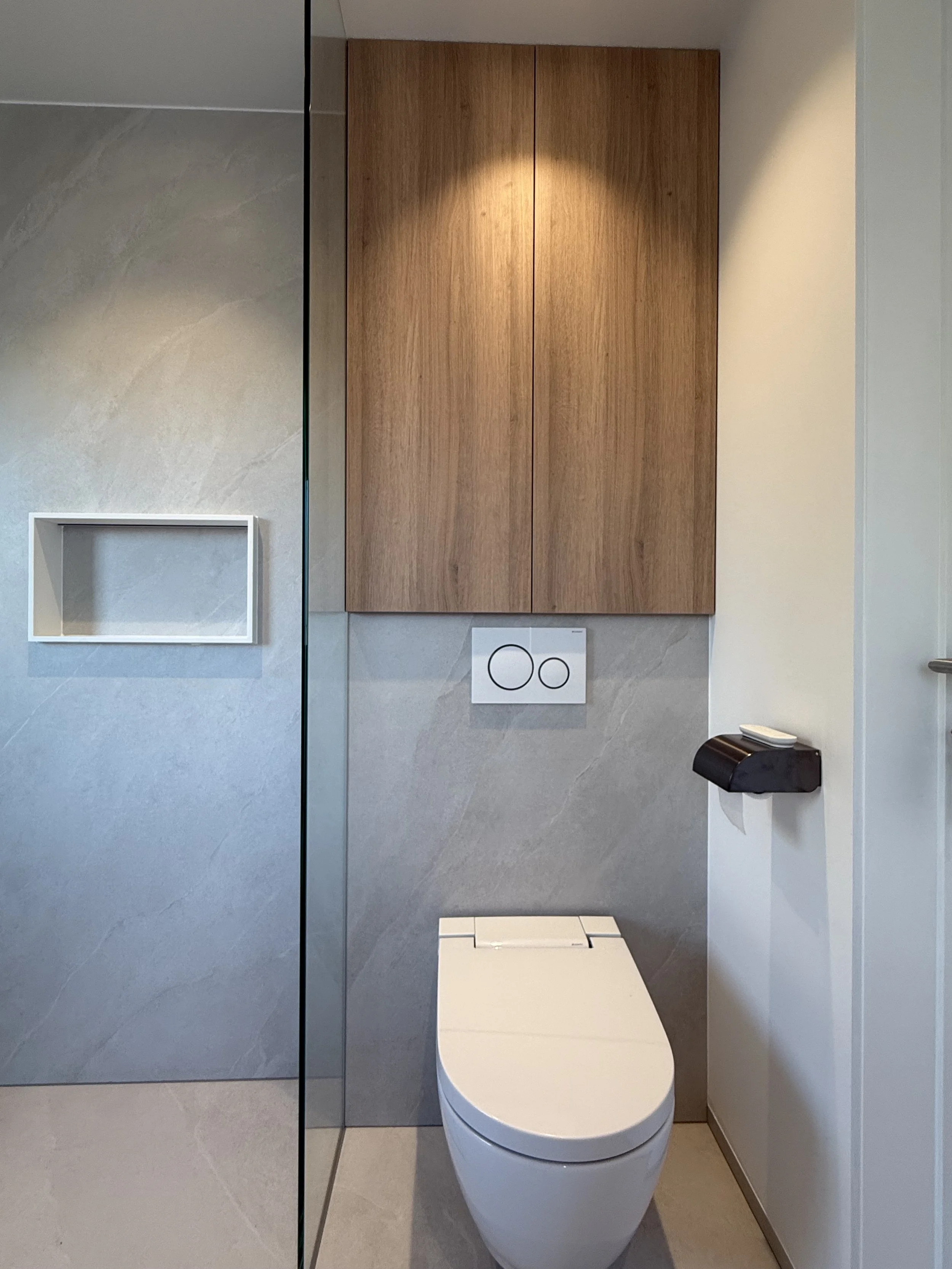 Moderne toilet in een badkamer met een houten kast boven het toilet, een wit toiletbril, en een inbouwspoelknop op de muur.