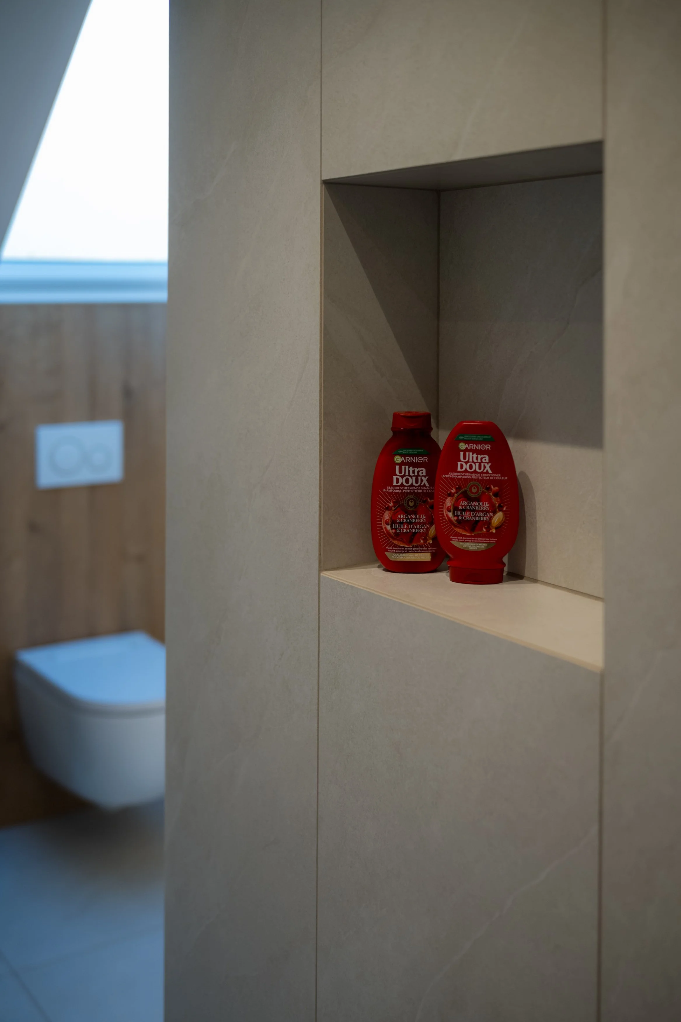 Twee flessen Garnier Ultra Doux shampoo en conditioner met arganolie en cranberry op een ingebouwde nis in een badkamerwand.