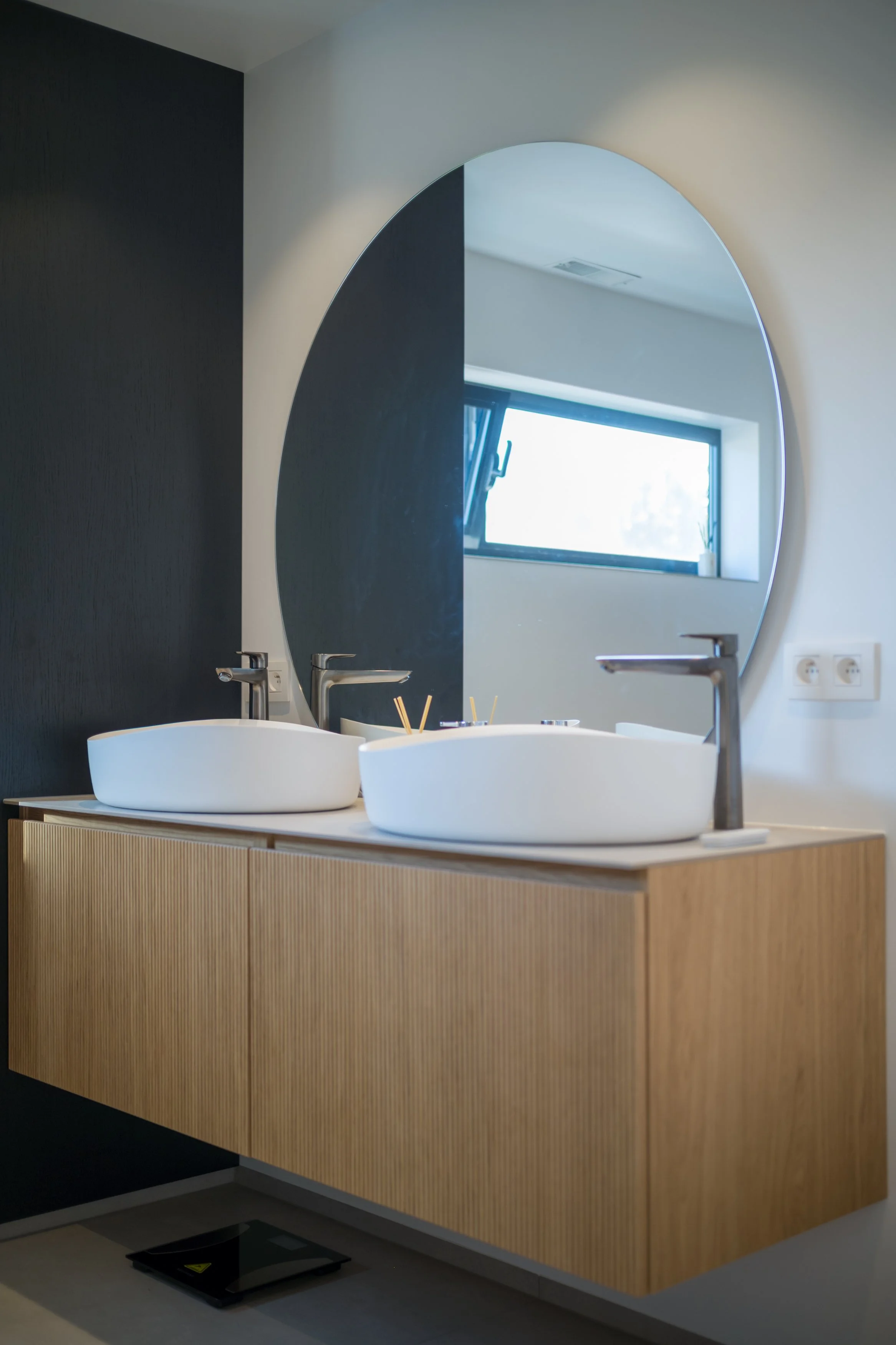 Een moderne badkamer met een houten wastafel, twee witte wasbakken en een grote ronde spiegel, naast een raampje.