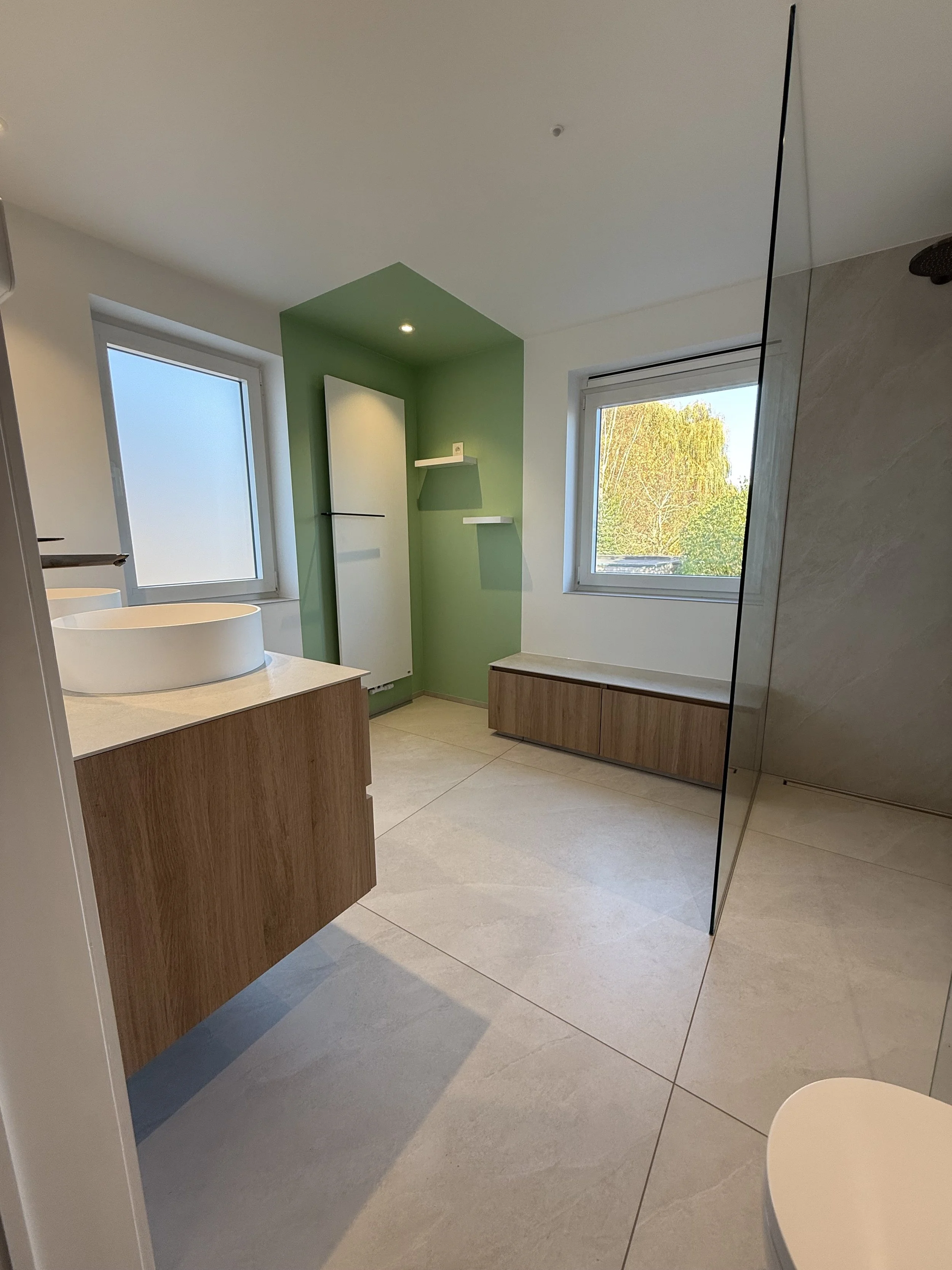 Moderne badkamer met groen accentmuur, houten laden, witte gootsteen, grote ramen met uitzicht op bomen, glazen scheidingswand en tegels op de vloer.