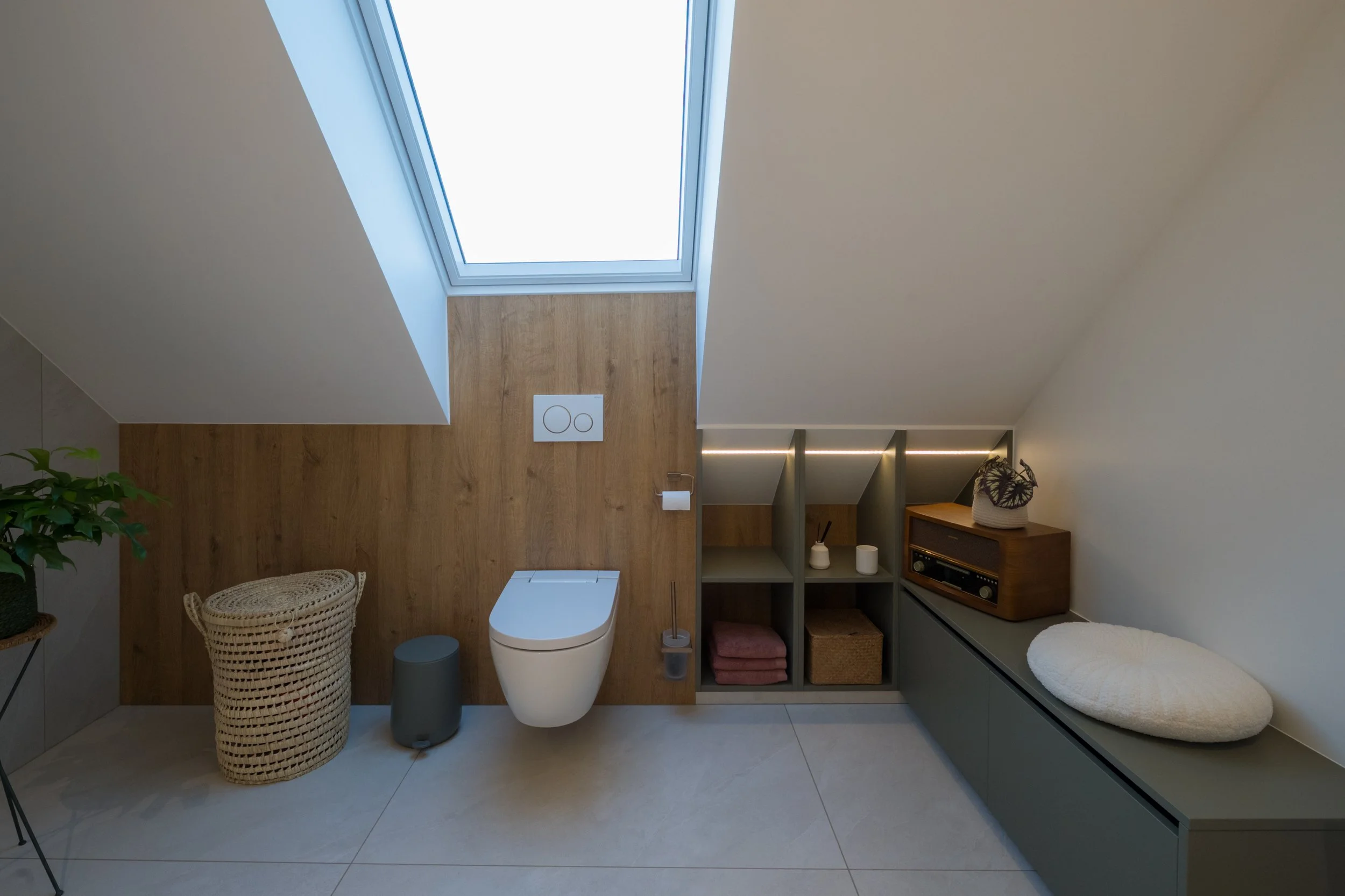 Moderne badkamer onder schuine gevel met een dakraam, toilet, open kast met handdoeken, planten, en een lage grijze opbergkast met decoratieve items.