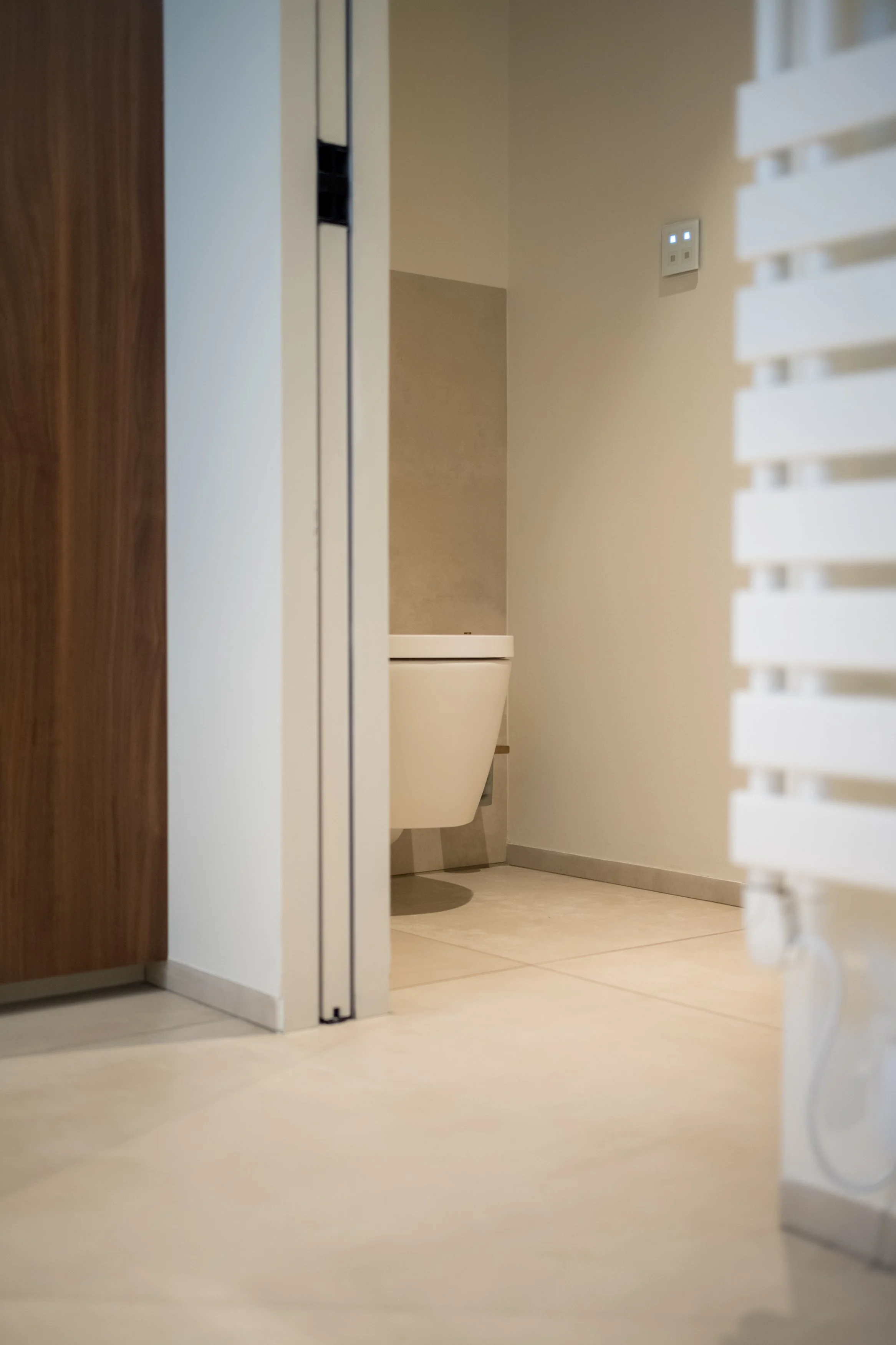 Kijk in een toiletruimte met een wandcloset toilet dat aan de muur hangt. Het is een nette, minimalistische badkamer met lichte tegels op de vloer en lichte muren. Rechts is een witte radiator zichtbaar.